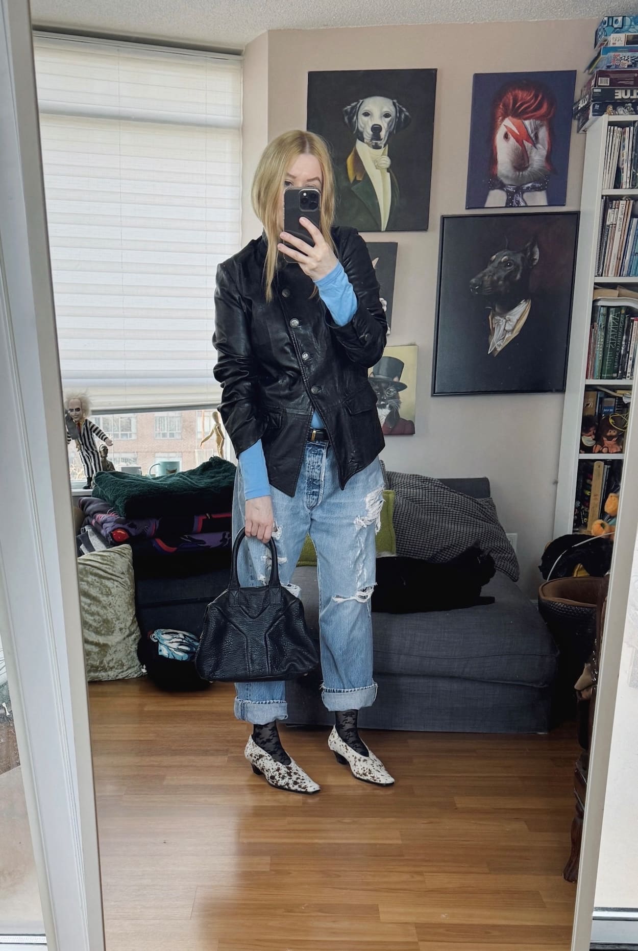 Sara of livelovesara is standing in front of a mirror taking a photo of her outfit. She is wearing a blue turtleneck under a vintage black leather jacket, vintage Levis, deer print heels, and a YSL Easy bag.