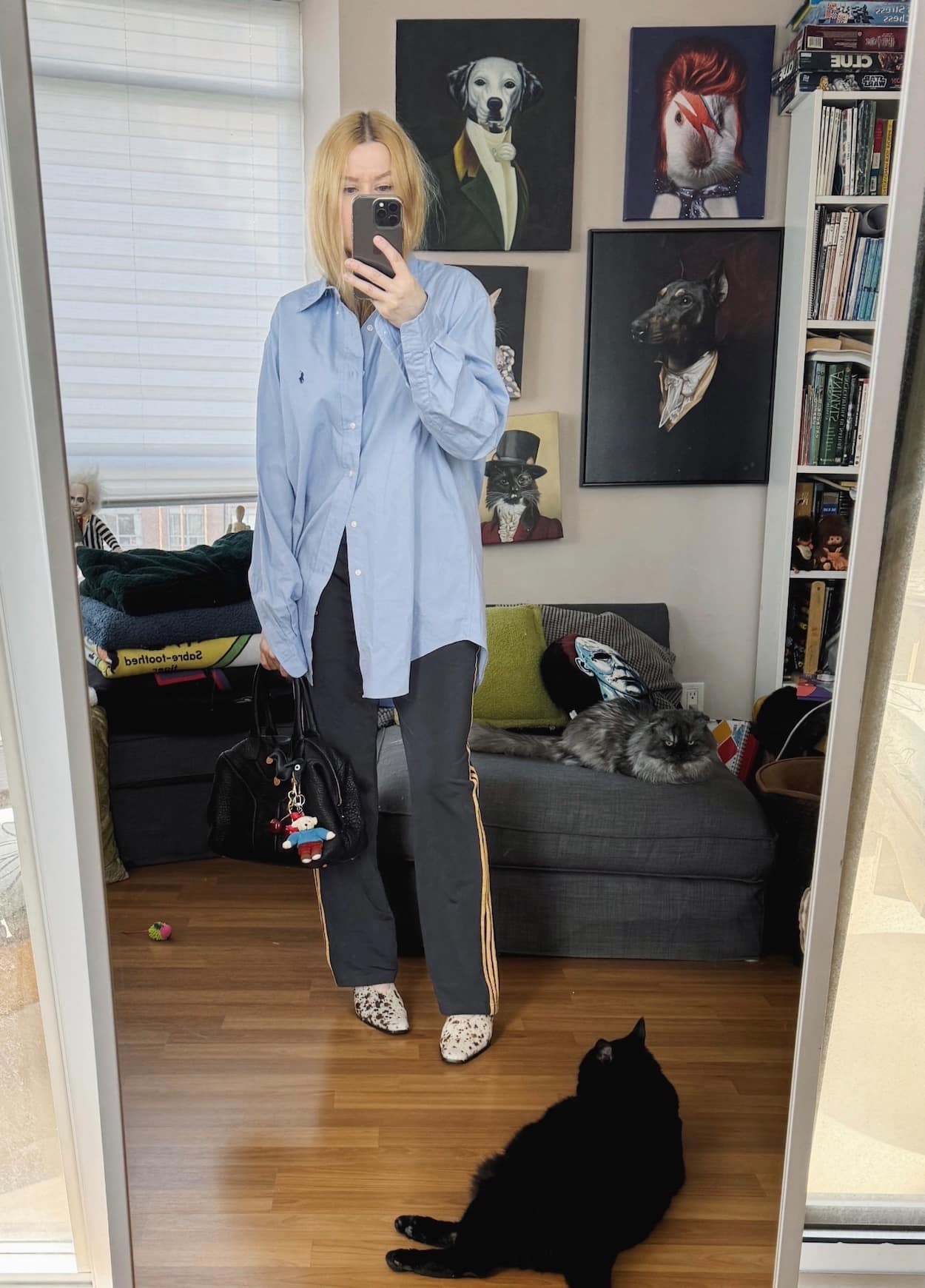 Sara of livelovesara is standing in front of a mirror taking a photo of her outfit. She is wearing a blue button up with black adidas track pants, cow print heels, and a YSL easy bag.
