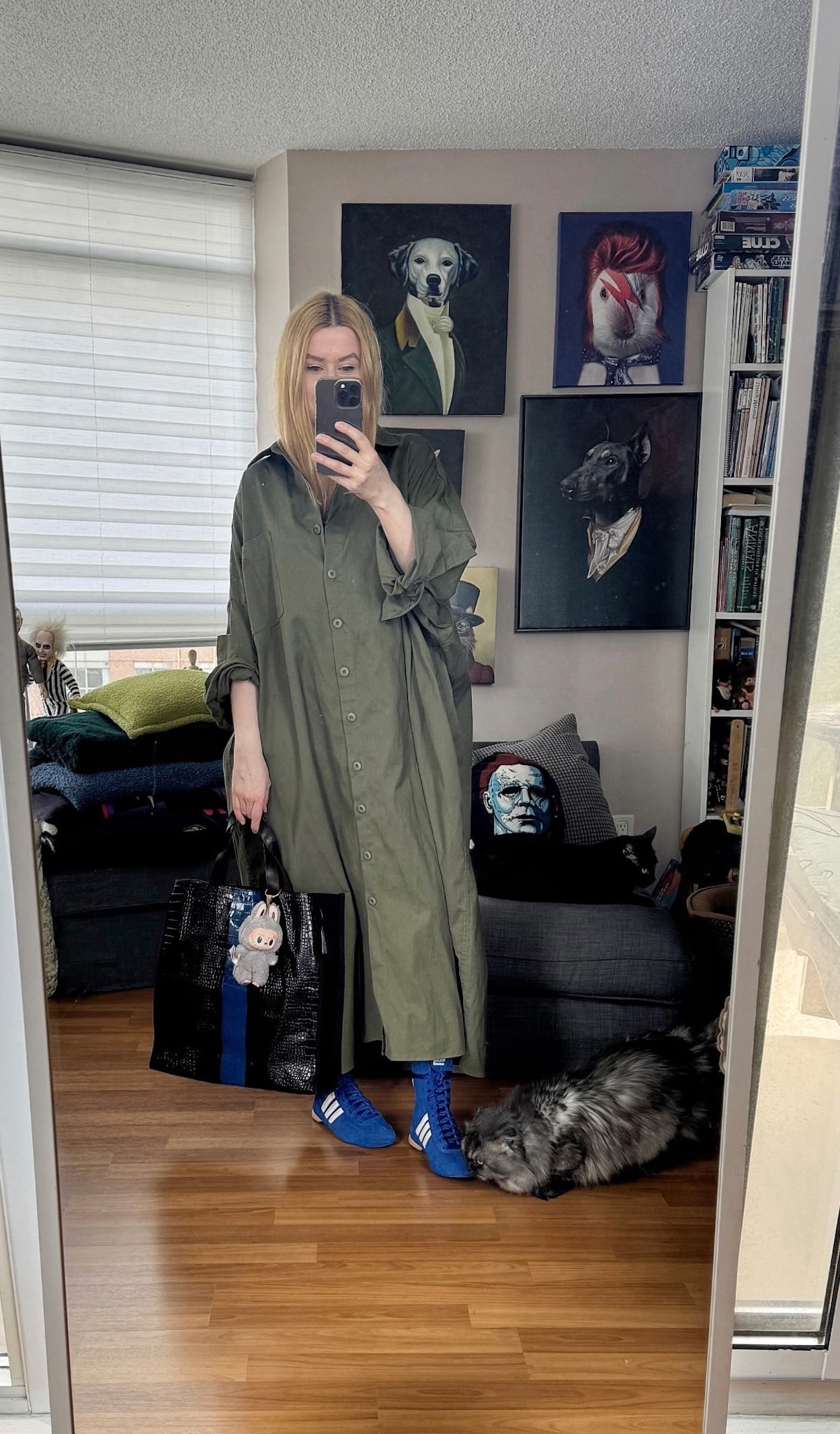 Sara of livelovesara is standing in front of a mirror taking a photo of her outfit. She is wearing an oversized green shirt dress, with blue Adidas Rasant high sneakers, and a striped tote.