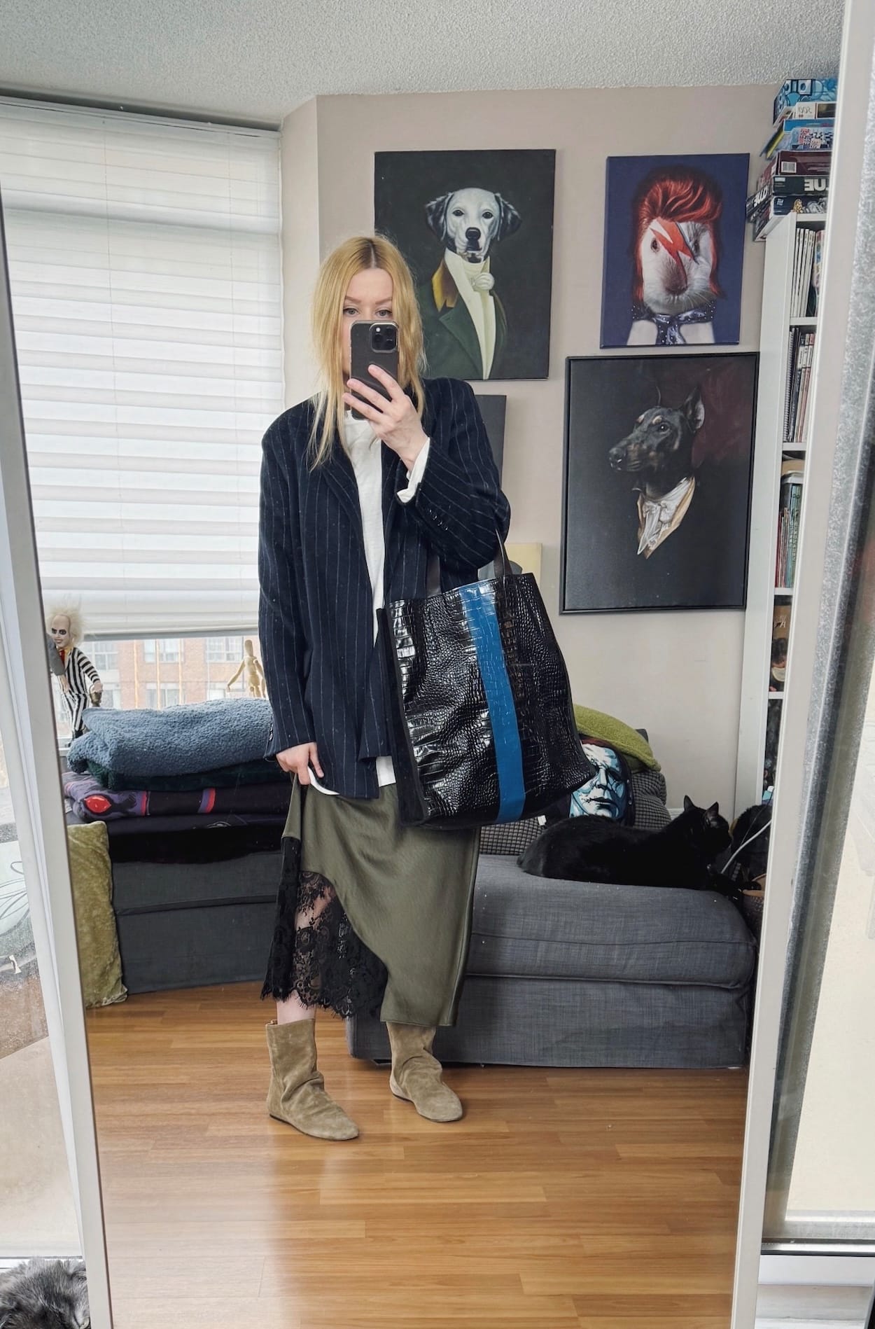 Sara of livelovesara is standing in front of a mirror taking a photo of her outfit. She is wearing a white long sleeve over a green slip dress, a striped blazer, suede boots, and is carrying a striped tote.