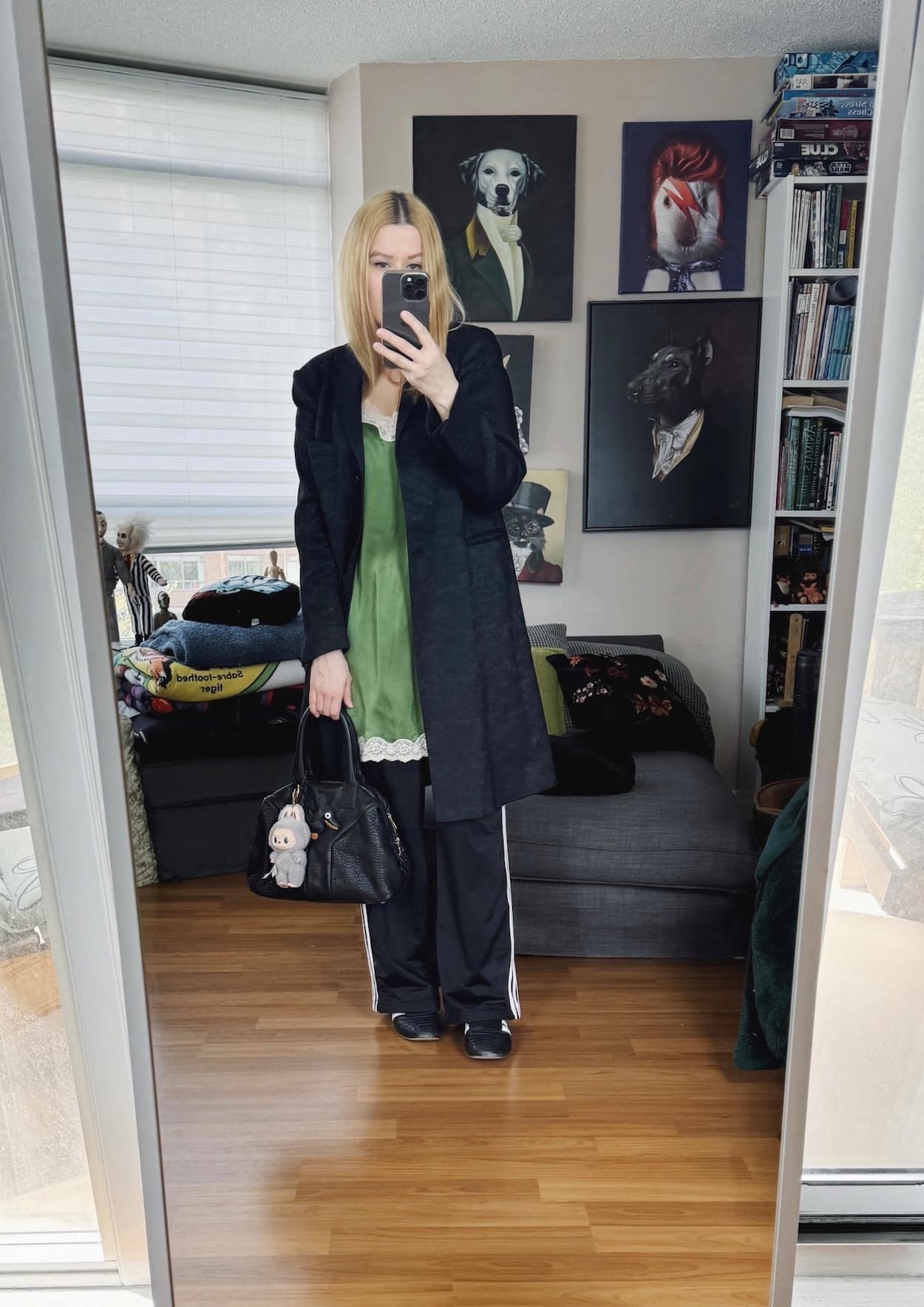 Sara of livelovesara is standing in front of a mirror taking a photo of her outfit. She is wearing a green camisole dress, adidas track pants, a kid length coat, and a ysl easy bag with a Labubu.