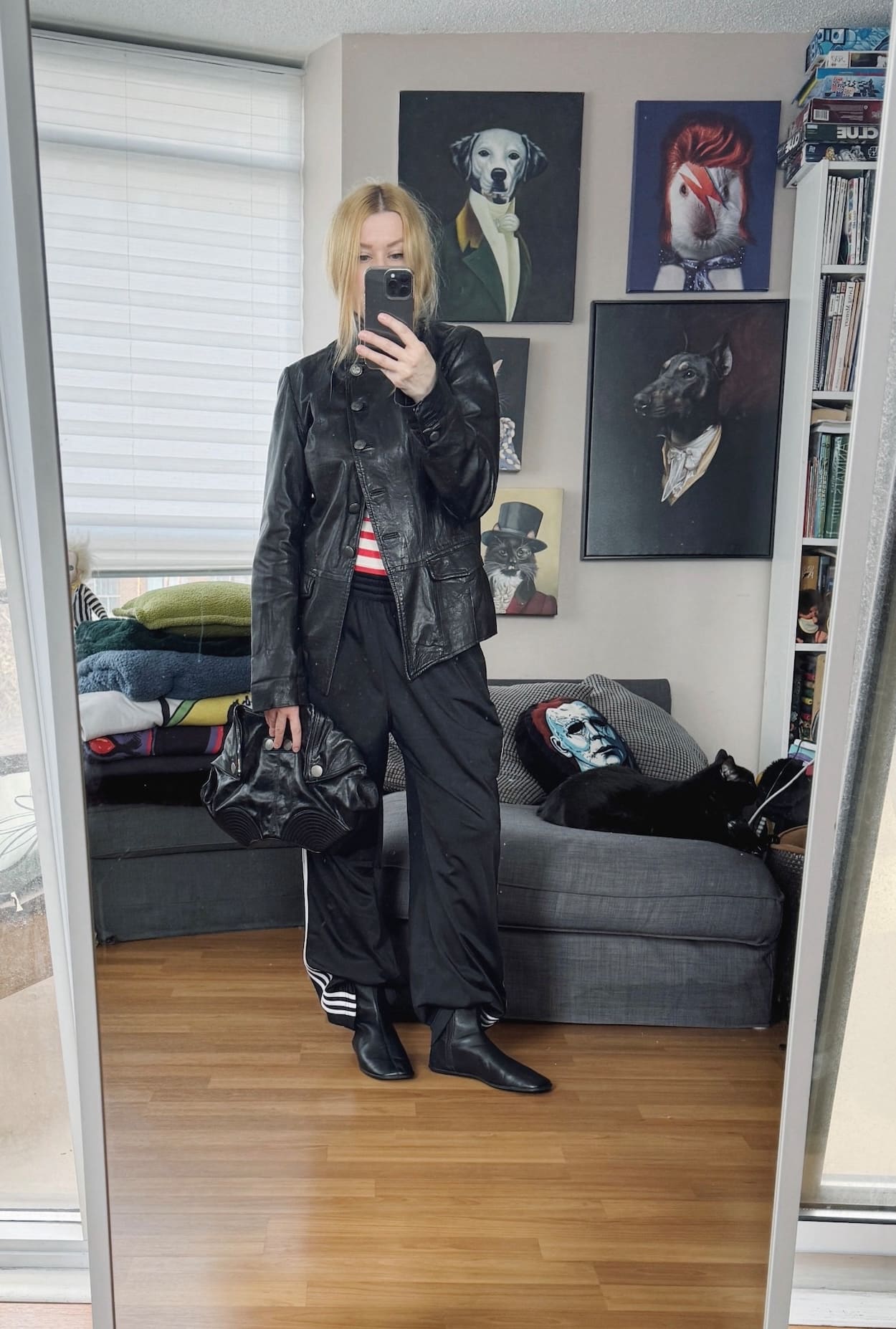 Sara of livelovesara is standing in front of a mirror taking a photo of her outfit. She is wearing a striped tank top under a vintage leather jacket, track pants, boots, and is carrying a clutch.