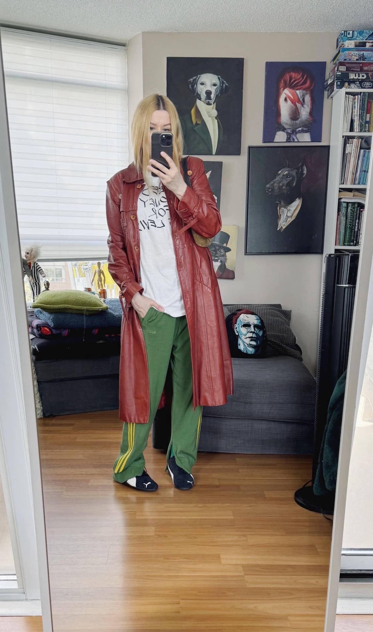 Sara of livelovesara is standing in front of a mirror taking a photo of her outfit. She is wearing a vintage tee with green Adidas track pants, a vintage leather trench, and a coach bag.