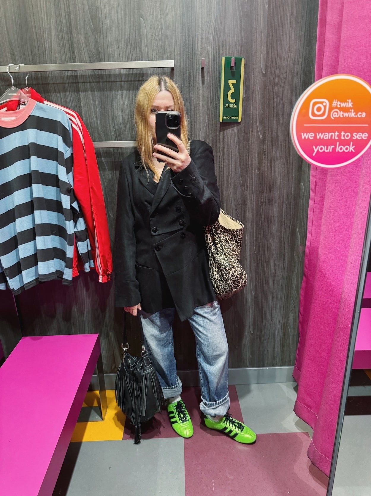 Sara of livelovesara is standing in front of a mirror taking a photo of her outfit. She is wearing a vintage blazer, vintage jeans, neon green adidas sneakers, and a vintage fringe bucket bag.