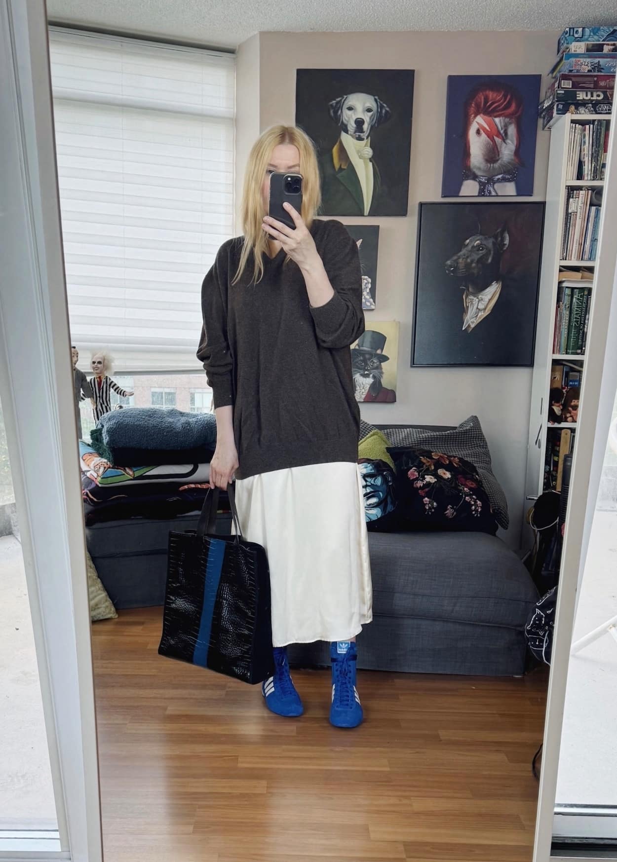 Sara of livelovesara is standing in front of a mirror taking a photo of her outfit. She is wearing a brown sweater over a cream slip dress, blue sneakers, and a tote.