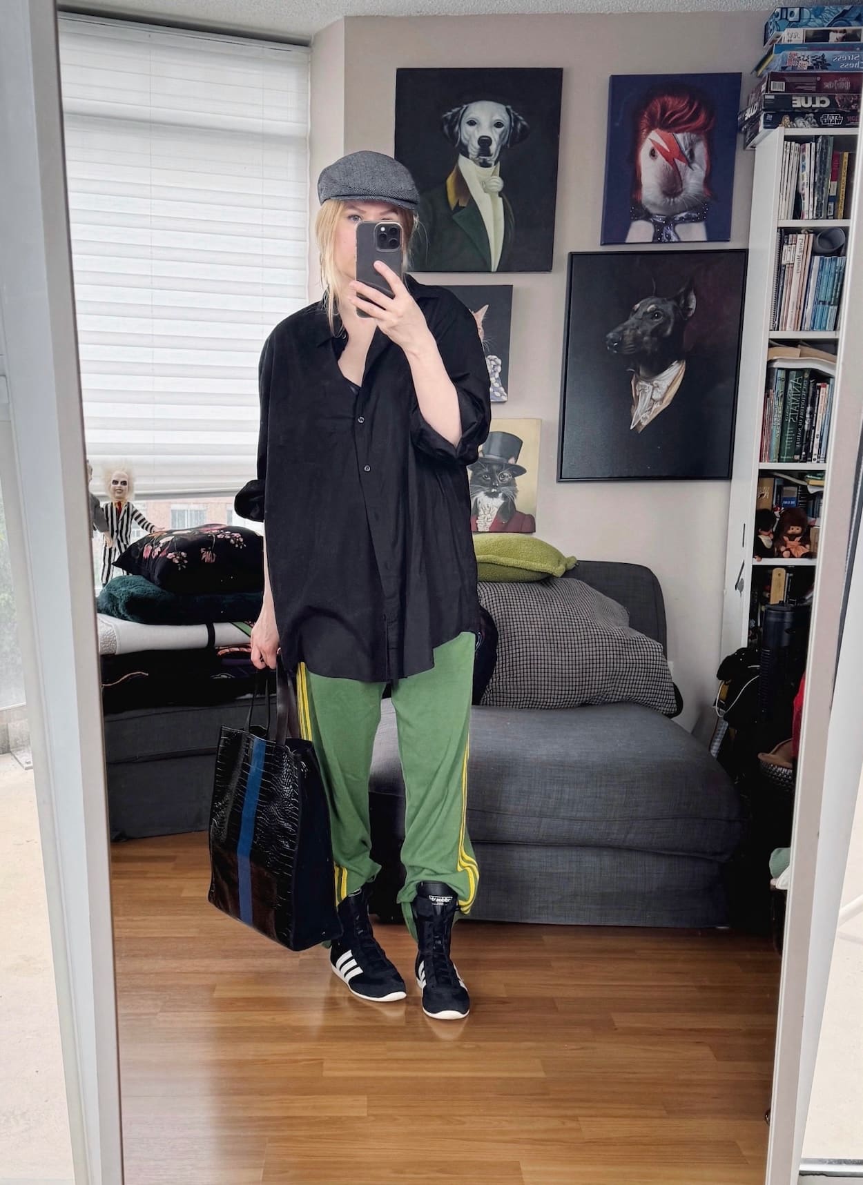Sara of livelovesara is standing in front of a mirror taking a photo of her outfit. She is wearing a black button up with green Adidas track pants, high adidas sneakers, a flat cap, and a tote.