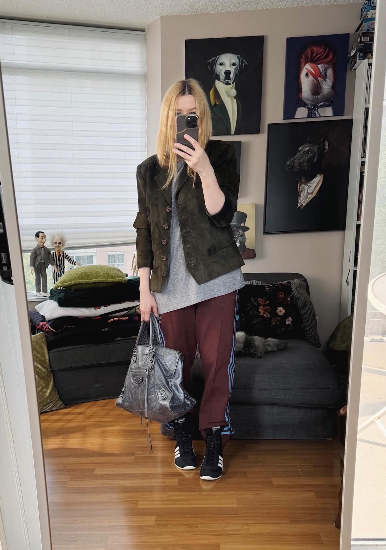 Sara of livelovesara is standing in front of a mirror taking a photo of her outfit. She is wearing a grey tee under an olive green suede jacket, adidas track pants, black sneakers, and a grey balenciaga bag.