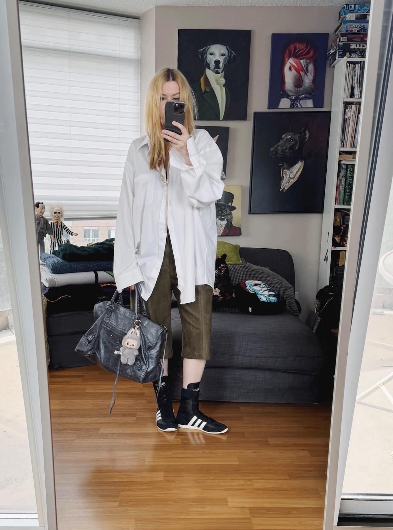Sara of livelovesara is standing in front of a mirror taking a photo of her outfit. She is wearing an oversized white button with green suede bermuda shirts, high adidas, and a balenciaga bag.