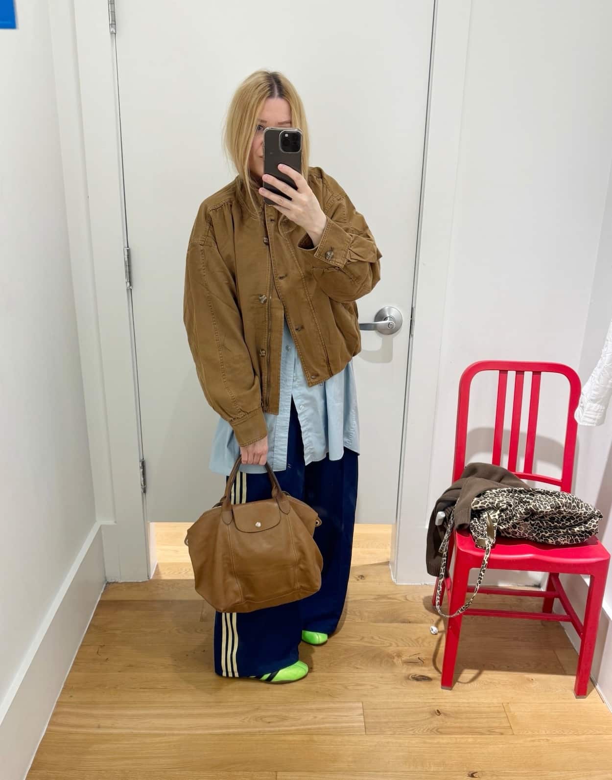 Sara of livelovesara is standing in front of a mirror taking a photo of her outfit. She is wearing an oversized blue button up with blue adidas track pants, neon green adidas sneakers, a brown jacket, and brown tote bag.