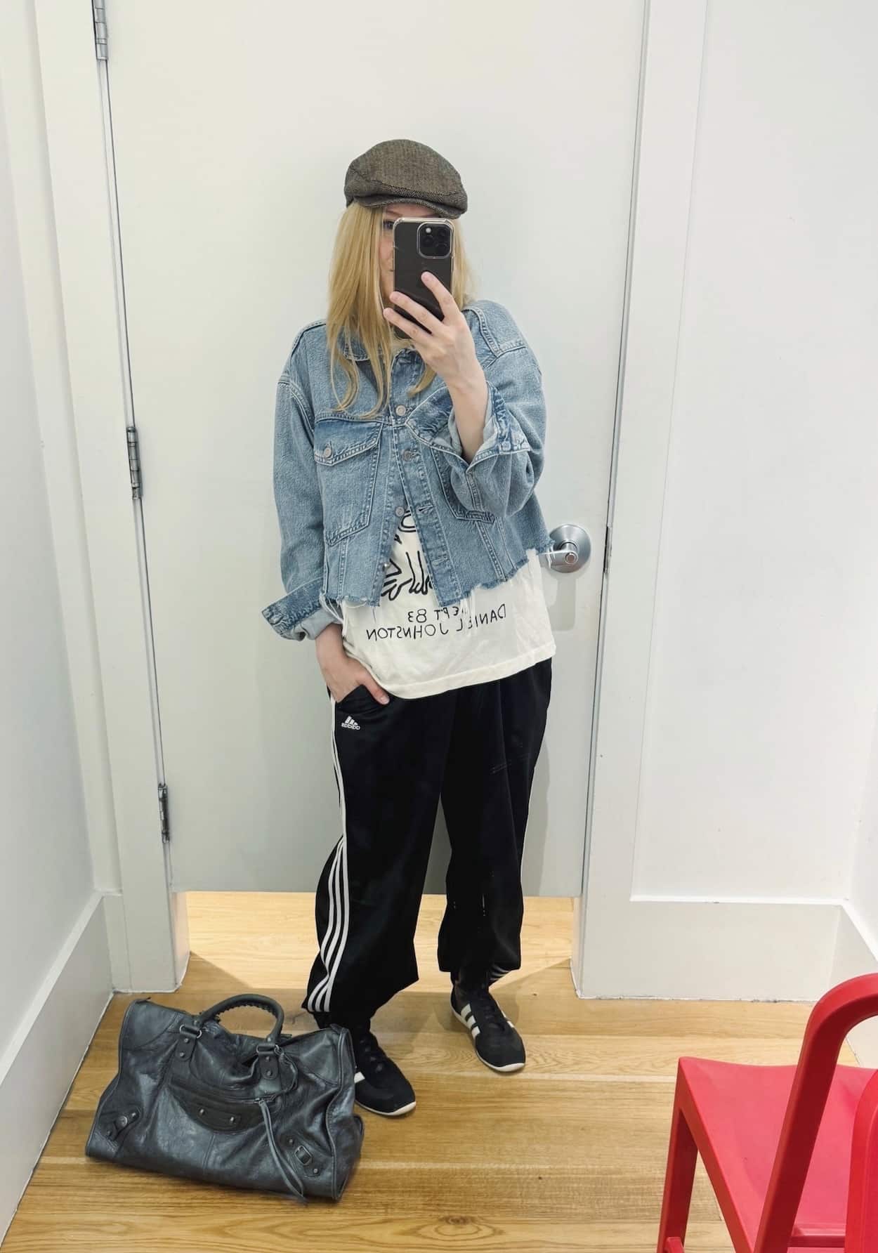Sara of livelovesara is standing in front of a mirror taking a photo of her outfit. She is wearing a white tee under a cropped denim jacket, adidas track pants, black high sneakers, a driving cap, and a grey bag.