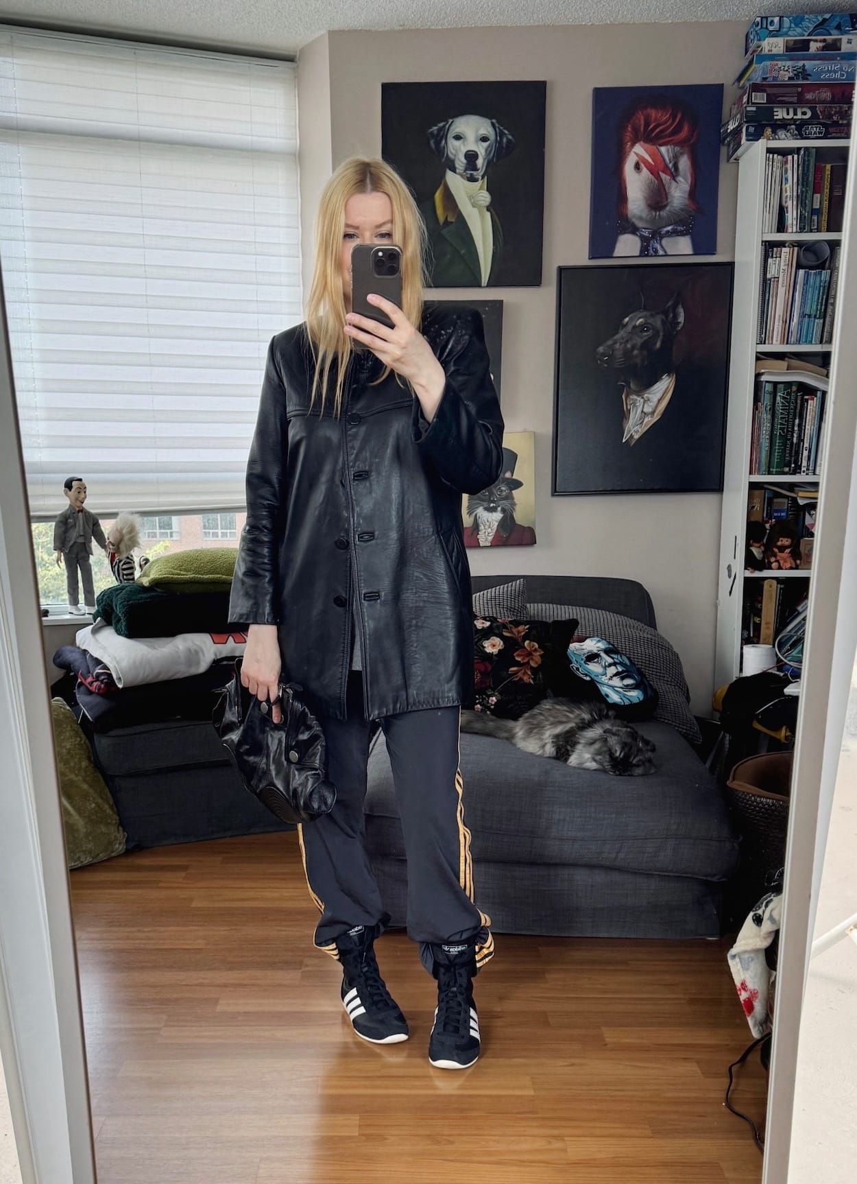 Sara of livelovesara is standing in front of a mirror taking a photo of her outfit. She is wearing a grey tee under a black leather jacket, black adidas track pants, adidas high sneakers, and a black clutch.