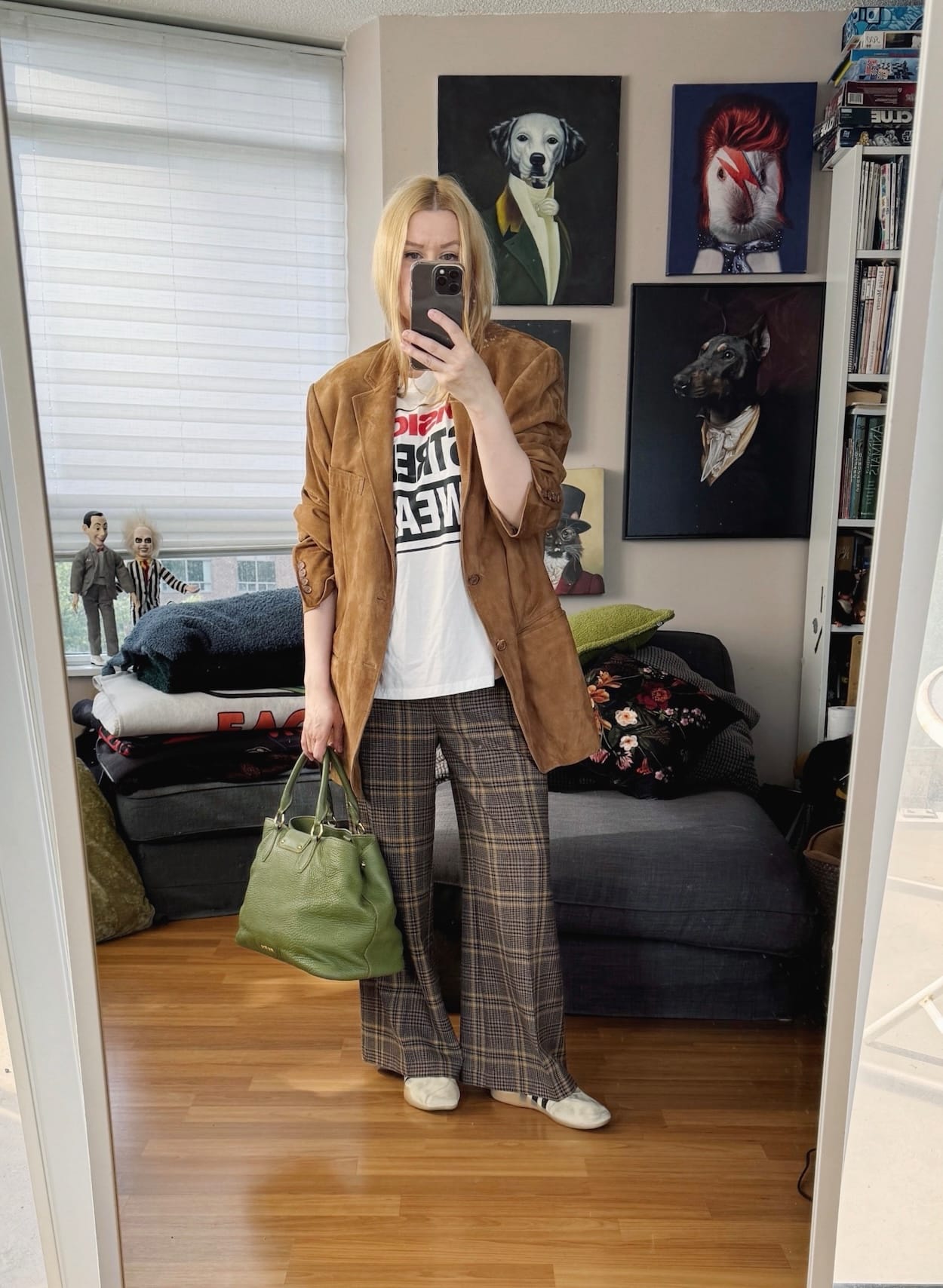 Sara of livelovesara is standing in front of a mirror taking a photo of her outfit. She is wearing a a vision streewear tee under a brown suede blazer, plaid pants, tan sneakers, and is carrying a green tote.