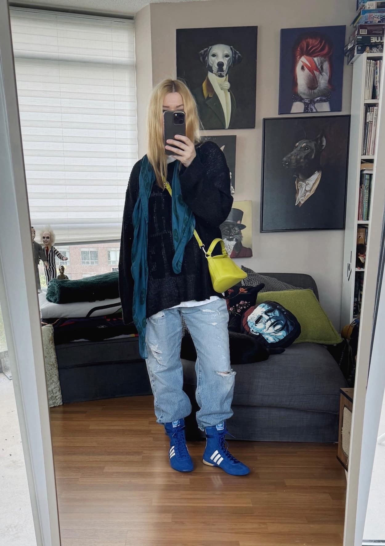 Sara of livelovesara is standing in front of a mirror taking a photo of her outfit. She is wearing a tee under an open weave sweater, vintage jeans, a scarf, sneakers, and a neon crossbody.