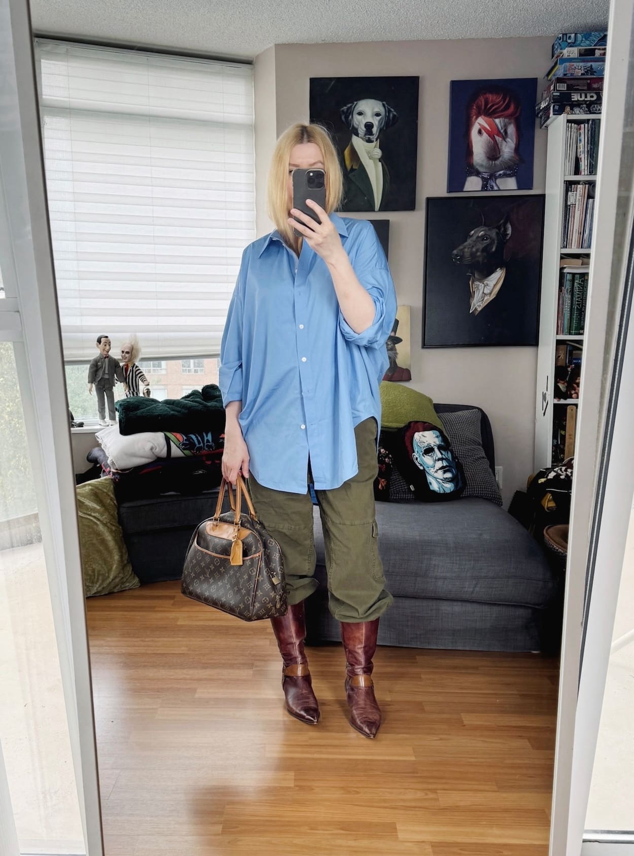 Sara of livelovesara is standing in front of a mirror taking a photo of her outfit. She is wearing a blue button up with cargos tucked into vintage boots, and is carrying a vintage louis vuitton bag.