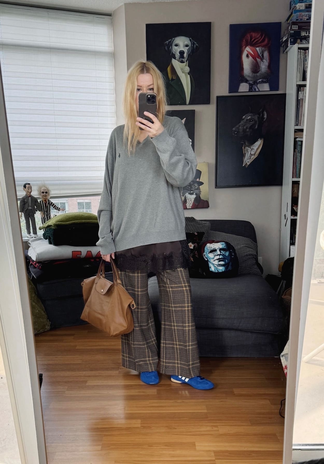 Sara of livelovesara is standing in front of a mirror taking a photo of her outfit. She is wearing a grey sweater over a camisole, plaid pants, blue sneakers, and is carrying a brown bag.