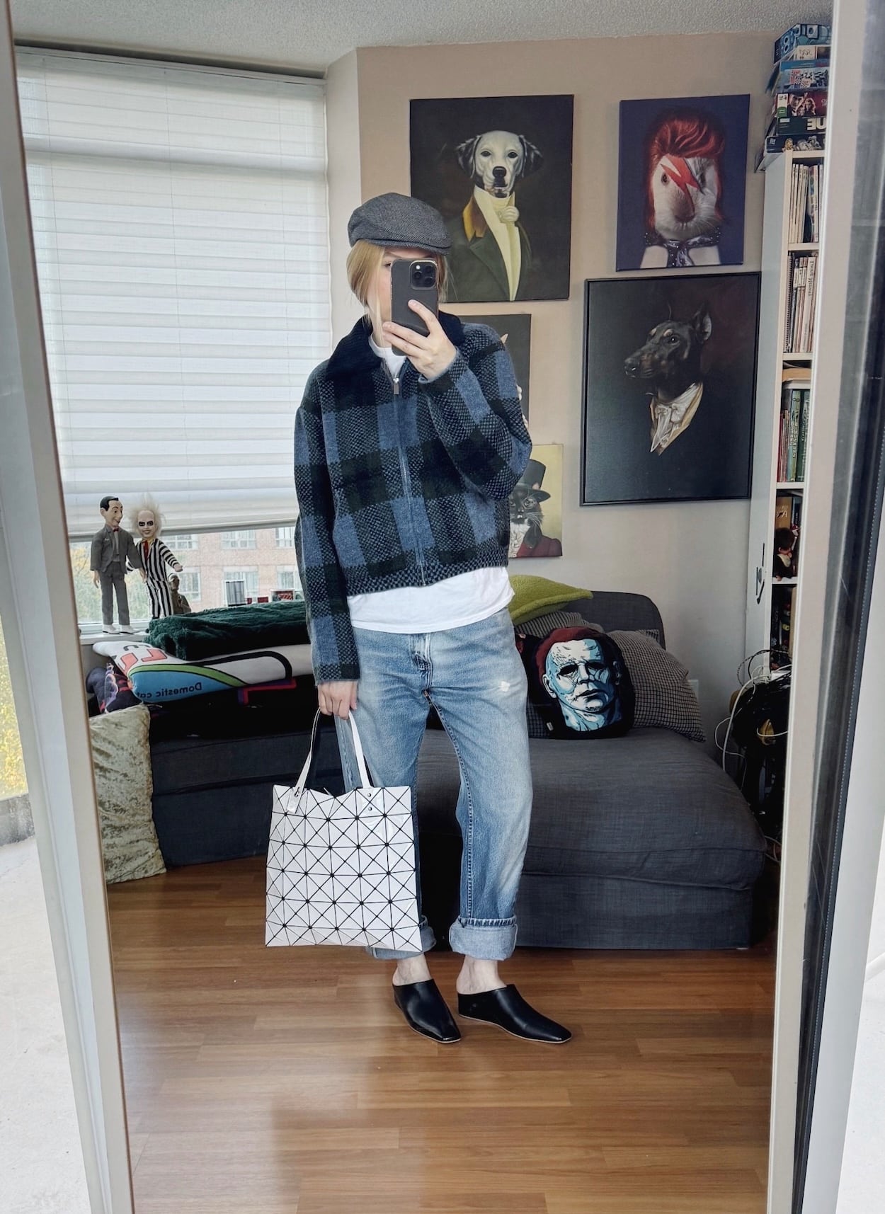Sara of livelovesara is standing in front of a mirror taking a photo of her outfit. She is wearing a blue plaid zip cardigan over a white t-shirt, vintage Levis, a driving cap, mules, and is carrying an Issey Miyake Bao Bao bag.