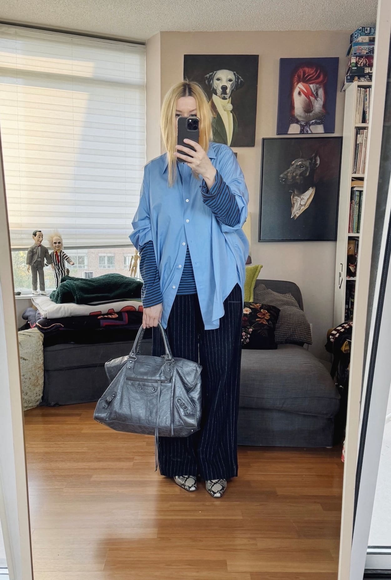 Sara of livelovesara is standing in front of a mirror taking a photo of her outfit. She is wearing a blue striped long sleeve under a blue button up, navy blue wool trousers, snake print boots, and a large Balenciaga bag.