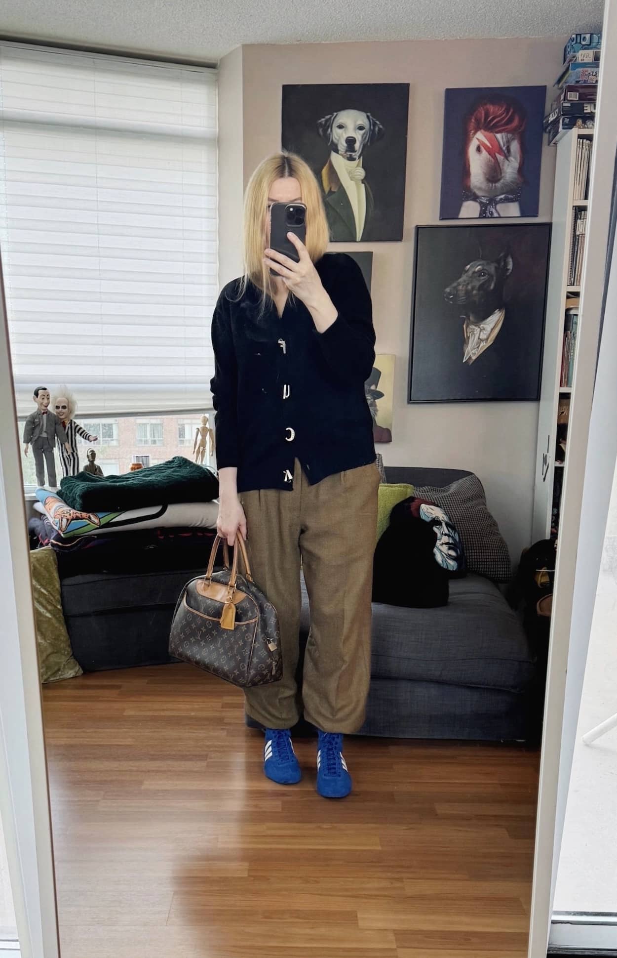 Sara of livelovesara is standing in front of a mirror taking a photo of her outfit. She is wearing a cardigan with F**K buttons, men's trousers, blue Adidas, and a vintage Louis Vuitton bag.