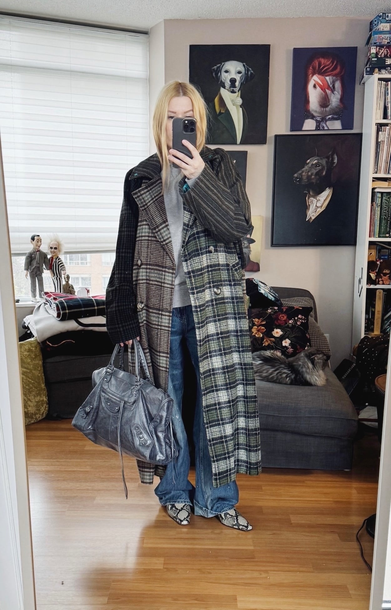 Sara of livelovesara is standing in front of a mirror taking a photo of her outfit. She is wearing a grey sweatshirt, paint splattered jeans, a mixed plaid coat, snake print boots, and a grey Balenciaga bag.