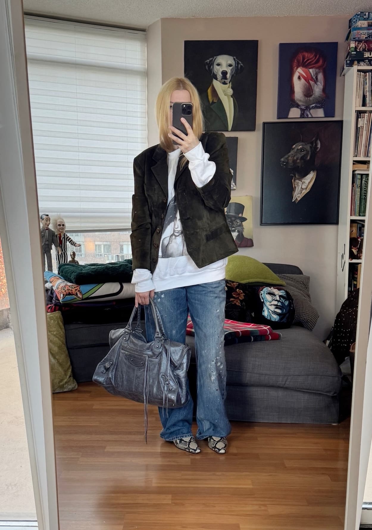 Sara of livelovesara is standing in front of a mirror taking a photo of her outfit. She is wearing a PJ Harvey sweatshirt with a olive green vintage suede blazer, paint splattered jeans, snake print boots, and is carrying a grey Balenciaga bag.