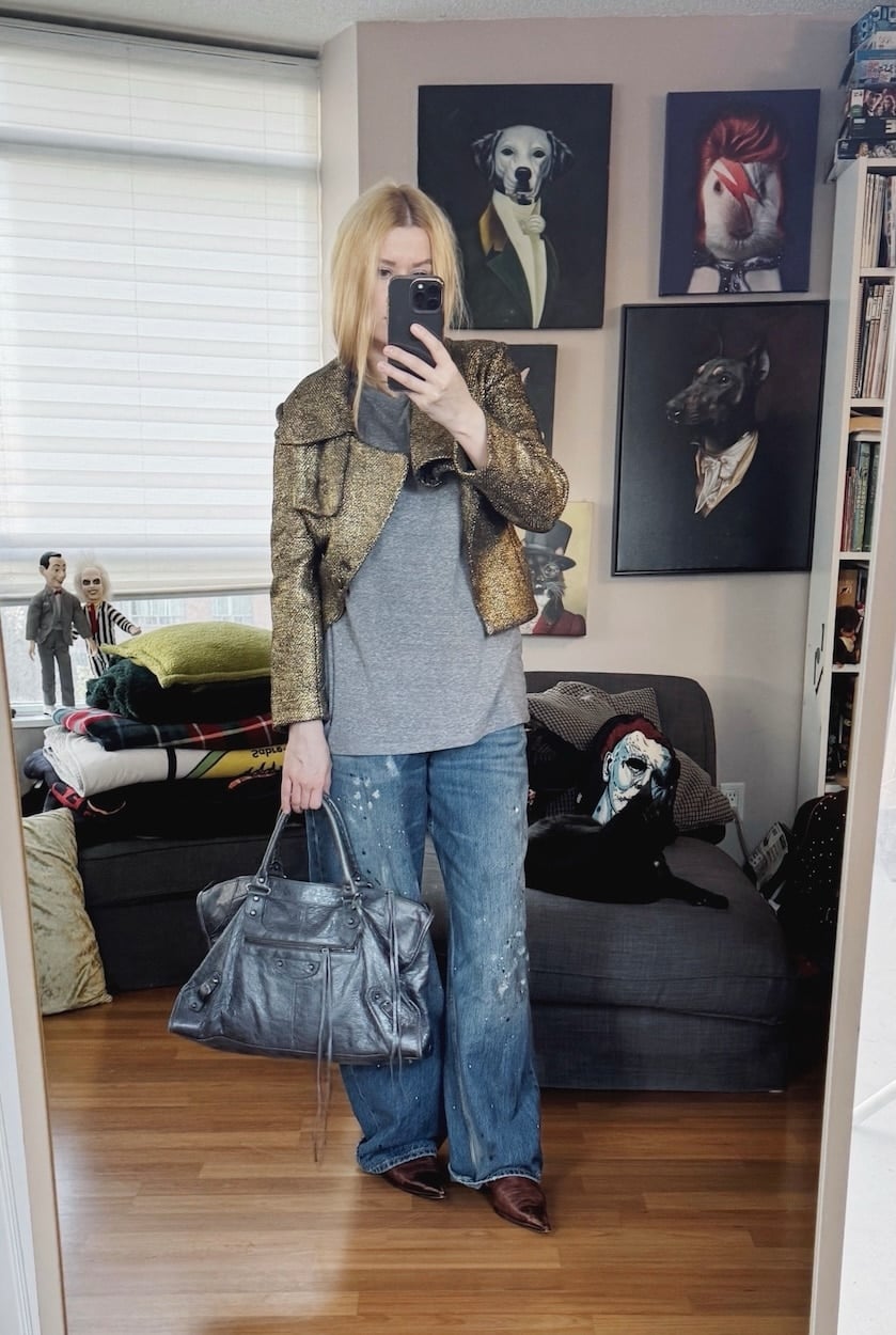 Sara of livelovesara is standing in front of a mirror taking a photo of her outfit. She is wearing a grey t-shirt under a gold cropped jacket, paint splattered jeans, brown vintage boots, and a grey Balenciaga city bag.