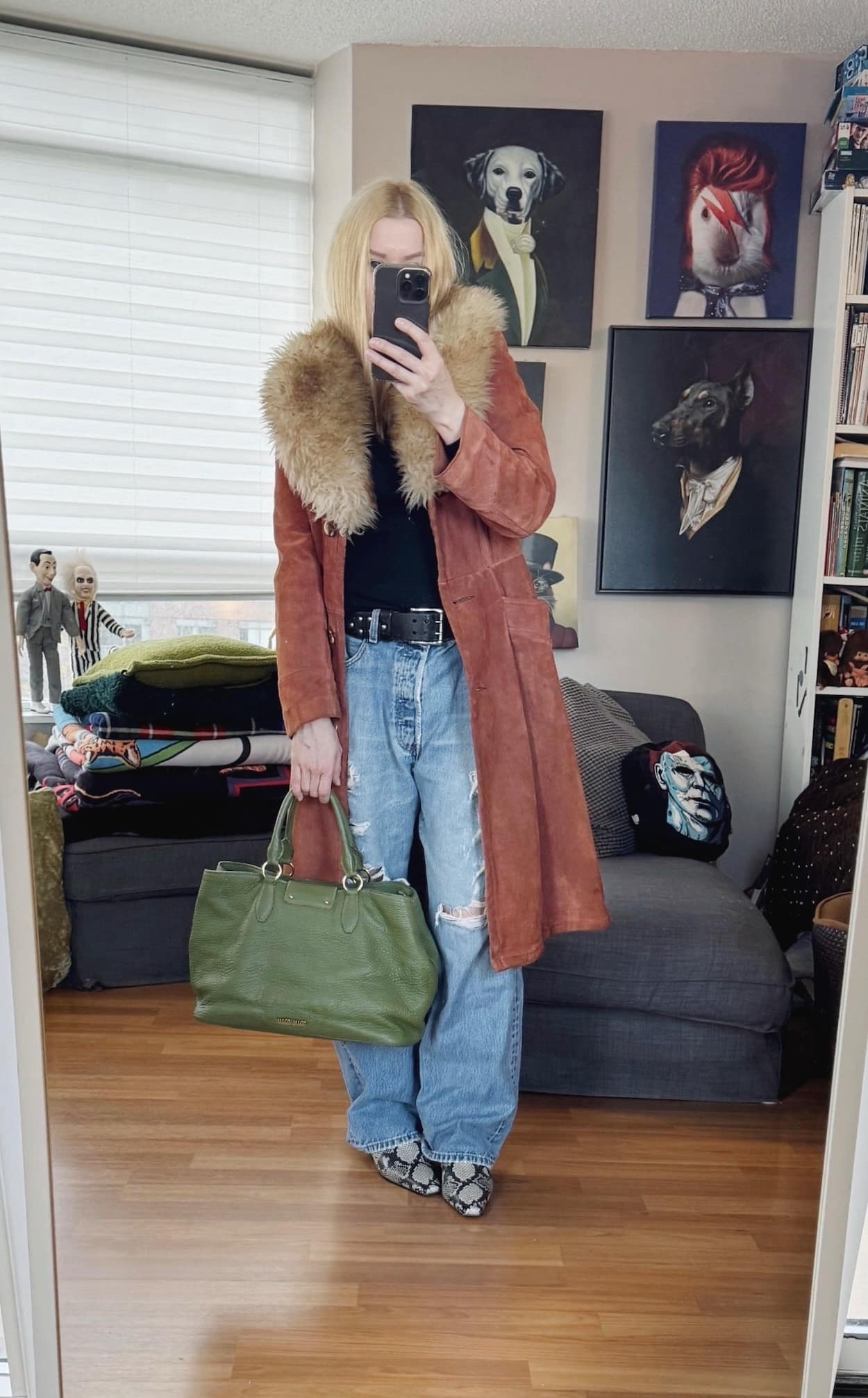 Sara of livelovesara is standing in front of a mirror taking a photo of her outfit. She is wearing a black turtleneck with vintage levis, a black studded belt, a 1970 suede trench with faux fur collar, and is carrying a green tote.