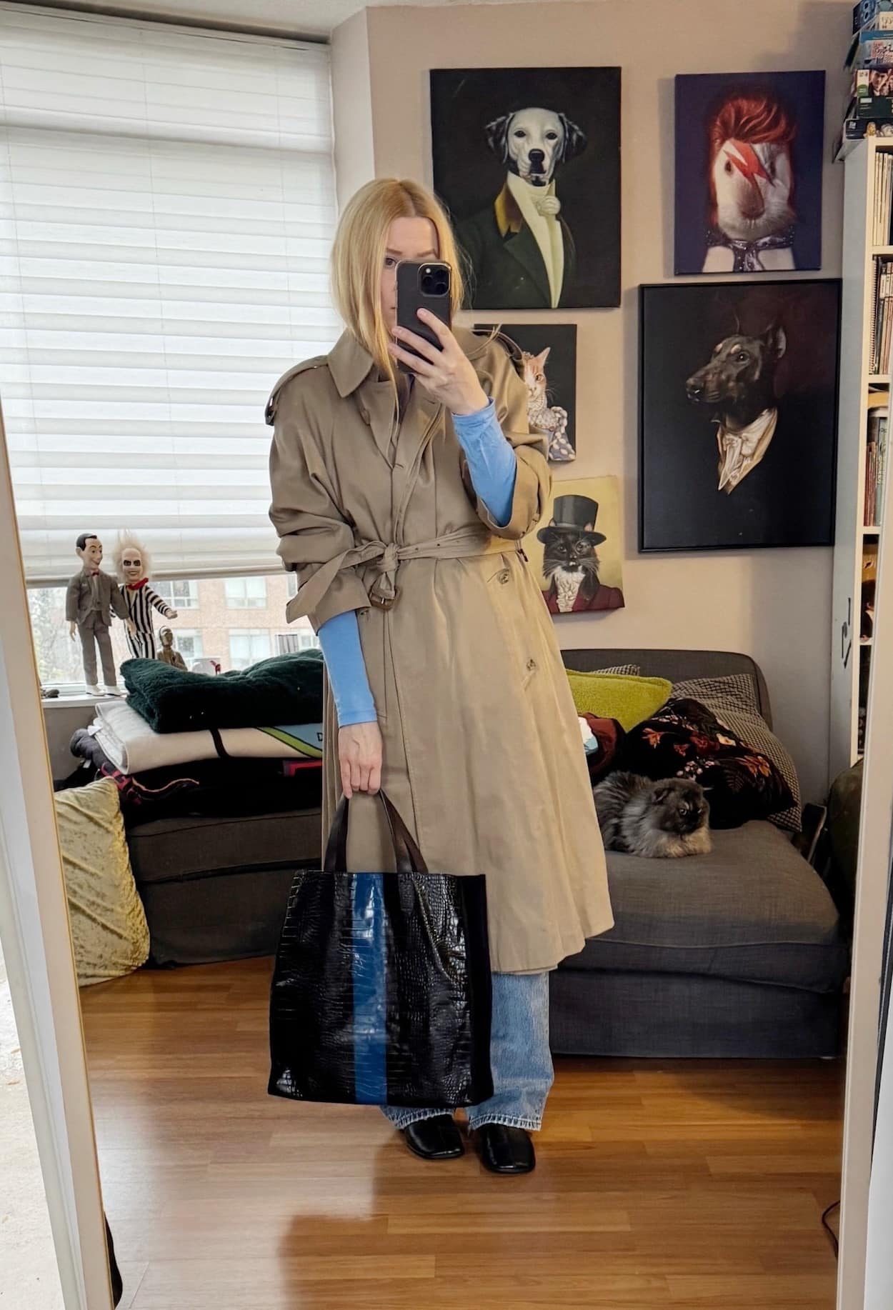 Sara of livelovesara is standing in front of a mirror taking a photo of her outfit. She is wearing a light blue turtleneck under a vintage trench, vintage levis, black flats boots, and is carrying a striped tote.
