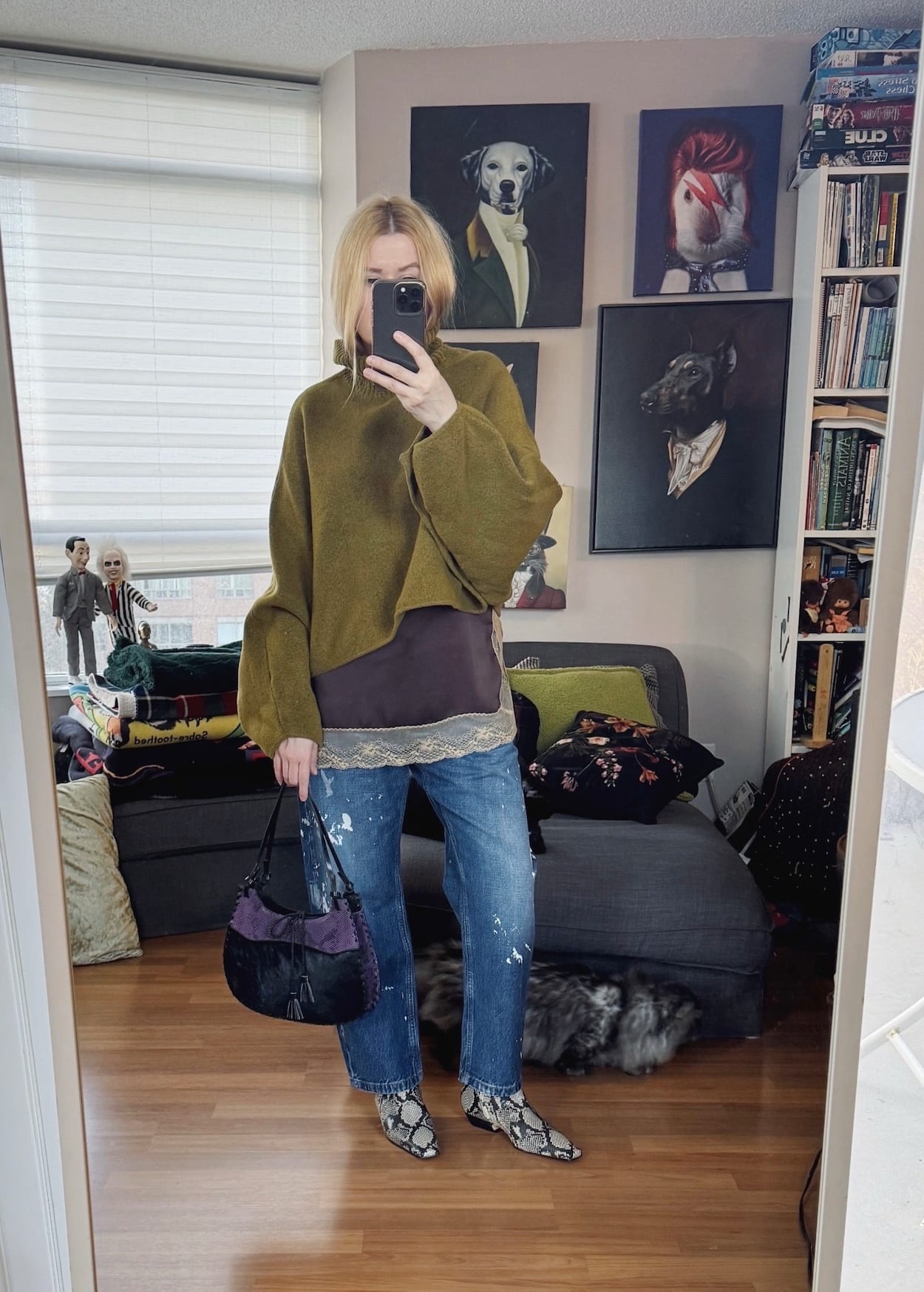Sara of livelovesara is standing in front of a mirror taking a photo of her outfit. She is wearing a brown camisole under a green turtleneck sweater, paint splattered jeans, snake print boots, and is carrying a purple and black vintage bag.