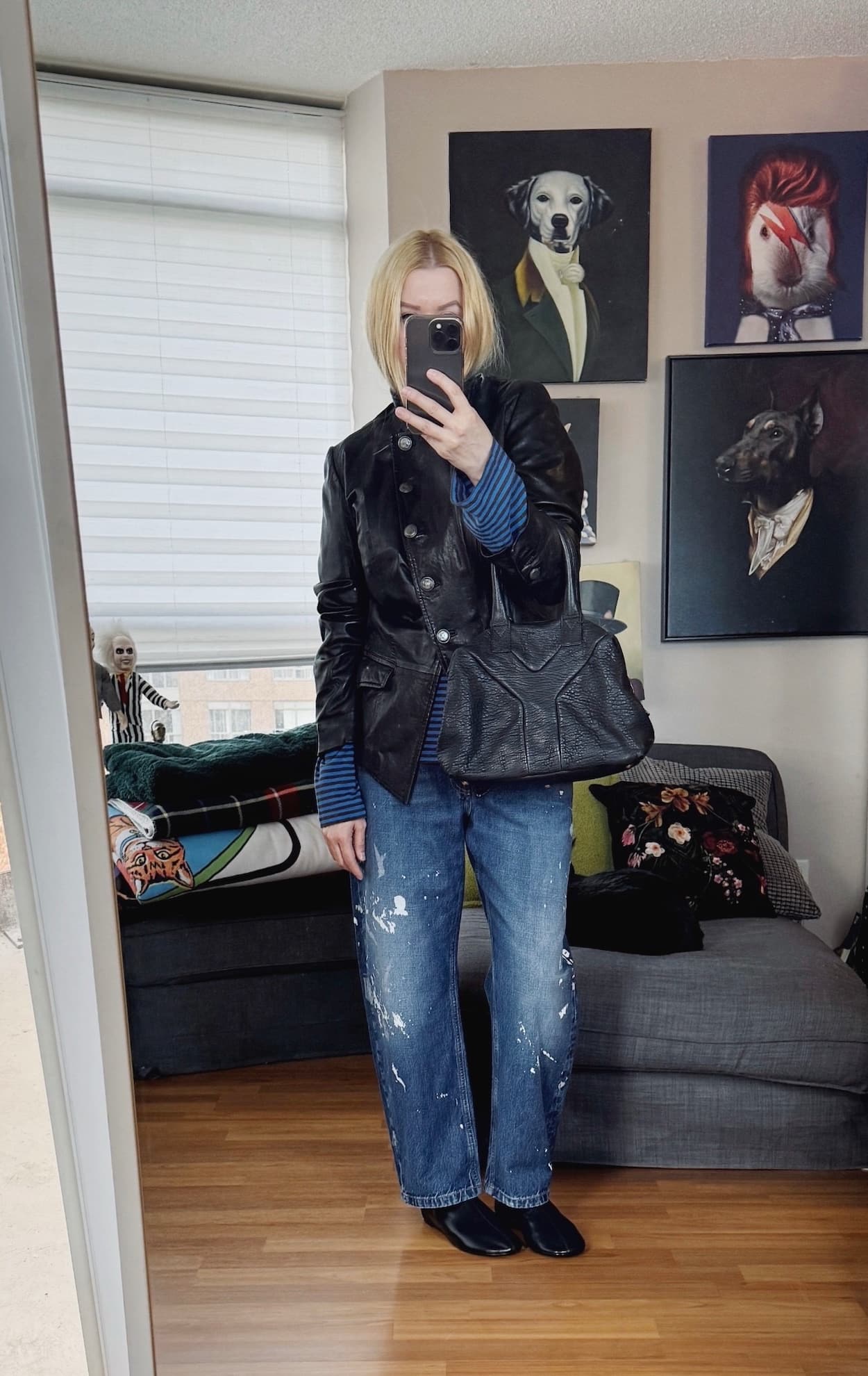Sara of livelovesara is standing in front of a mirror taking a photo of her outfit. She is wearing a blue striped long sleeve under a vintage leather jacket, paint splatered jeans, black boots, and is carrying a YSL easy bag.