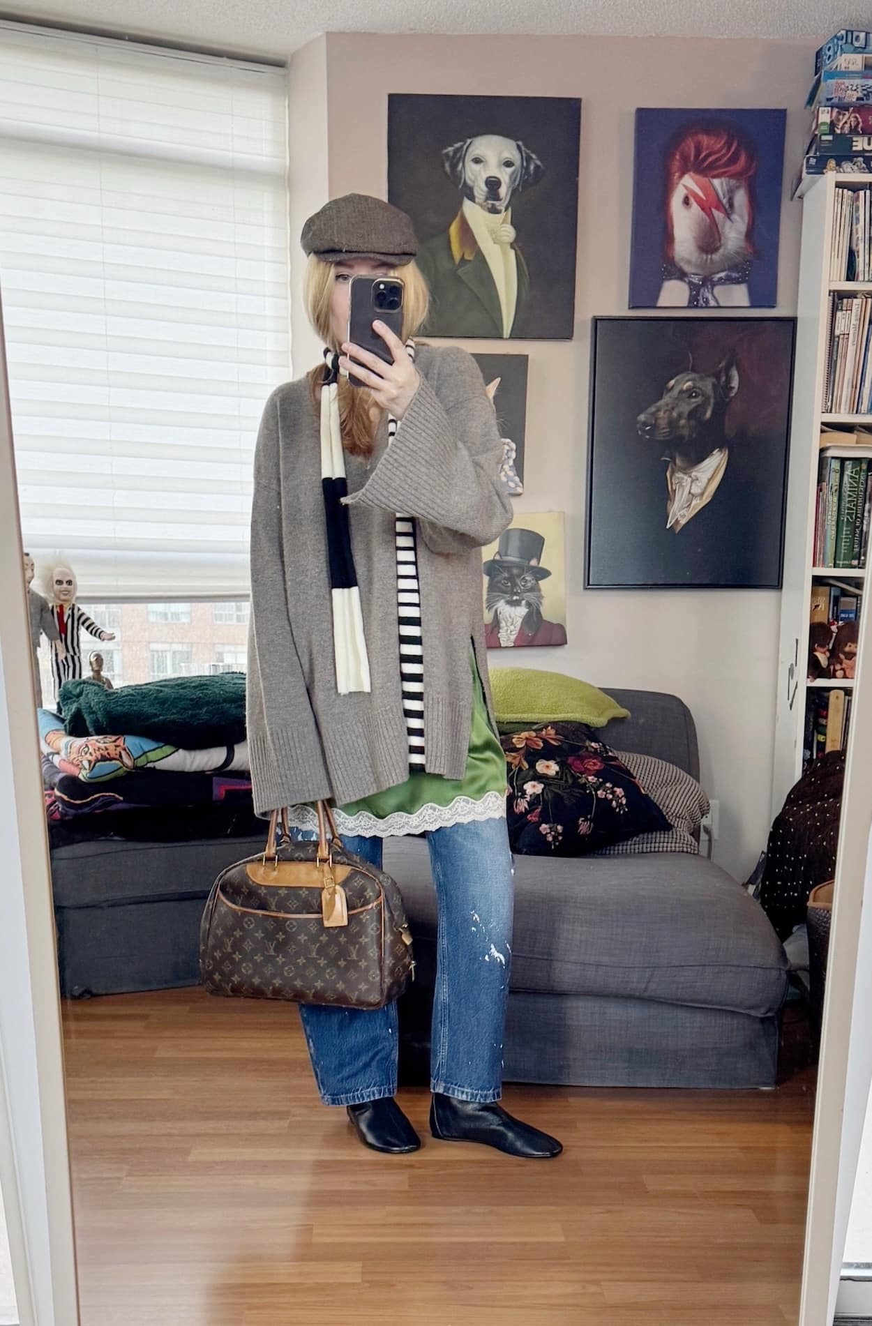 Sara of livelovesara is standing in front of a mirror taking a photo of her outfit. She is wearing an oversized sweater over a long green camisole, paint splatter jeans black boots, a skinny scarf, a driving cap, and is carrying a vintage Louis Vuitton bag.