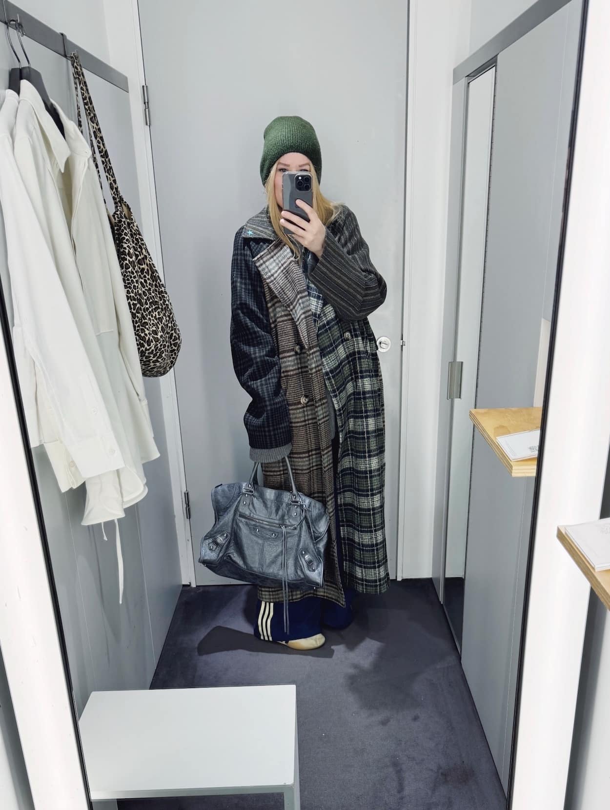Sara of livelovesara is standing in front of a mirror taking a photo of her outfit. She is wearing a grey sweater and blue Adidas track pants under a long mixed plaid coat, a green beanie, Adidas sneakers, and a large Balenciaga bag.