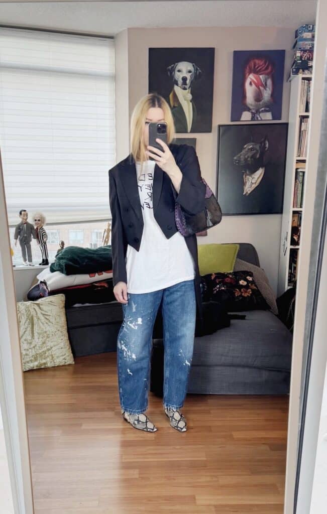 Sara of livelovesara is standing in front of a mirror taking a photo of her outfit. She is wearing a vintage tee under a vintage tailcoat, paint splatter jeans, snake print boots, and a vintage bag.