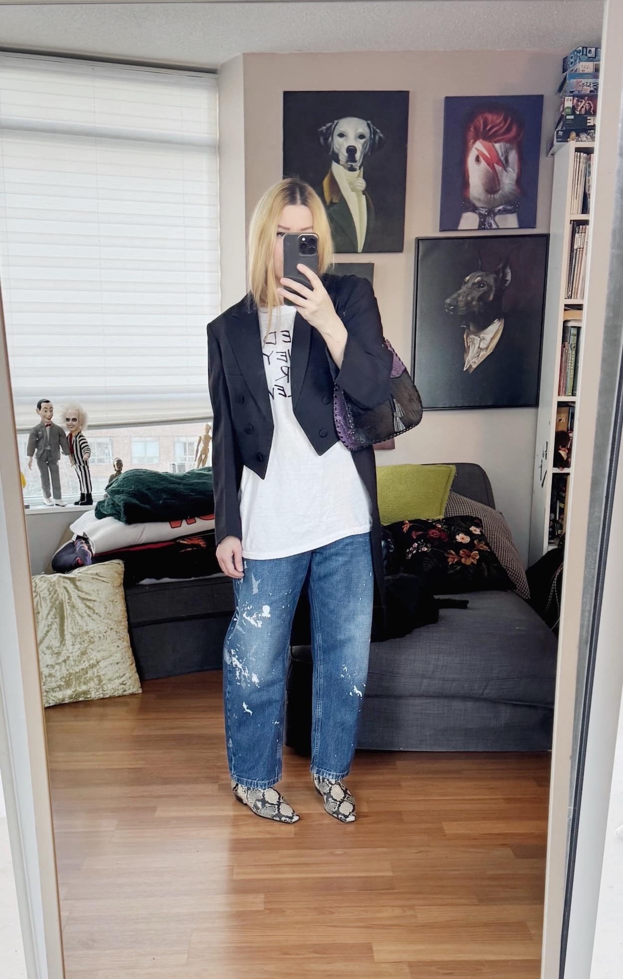 Sara of livelovesara is standing in front of a mirror taking a photo of her outfit. She is wearing a vintage tee under a vintage tailcoat, paint splatter jeans, snaker print boots, and a vintage bag.