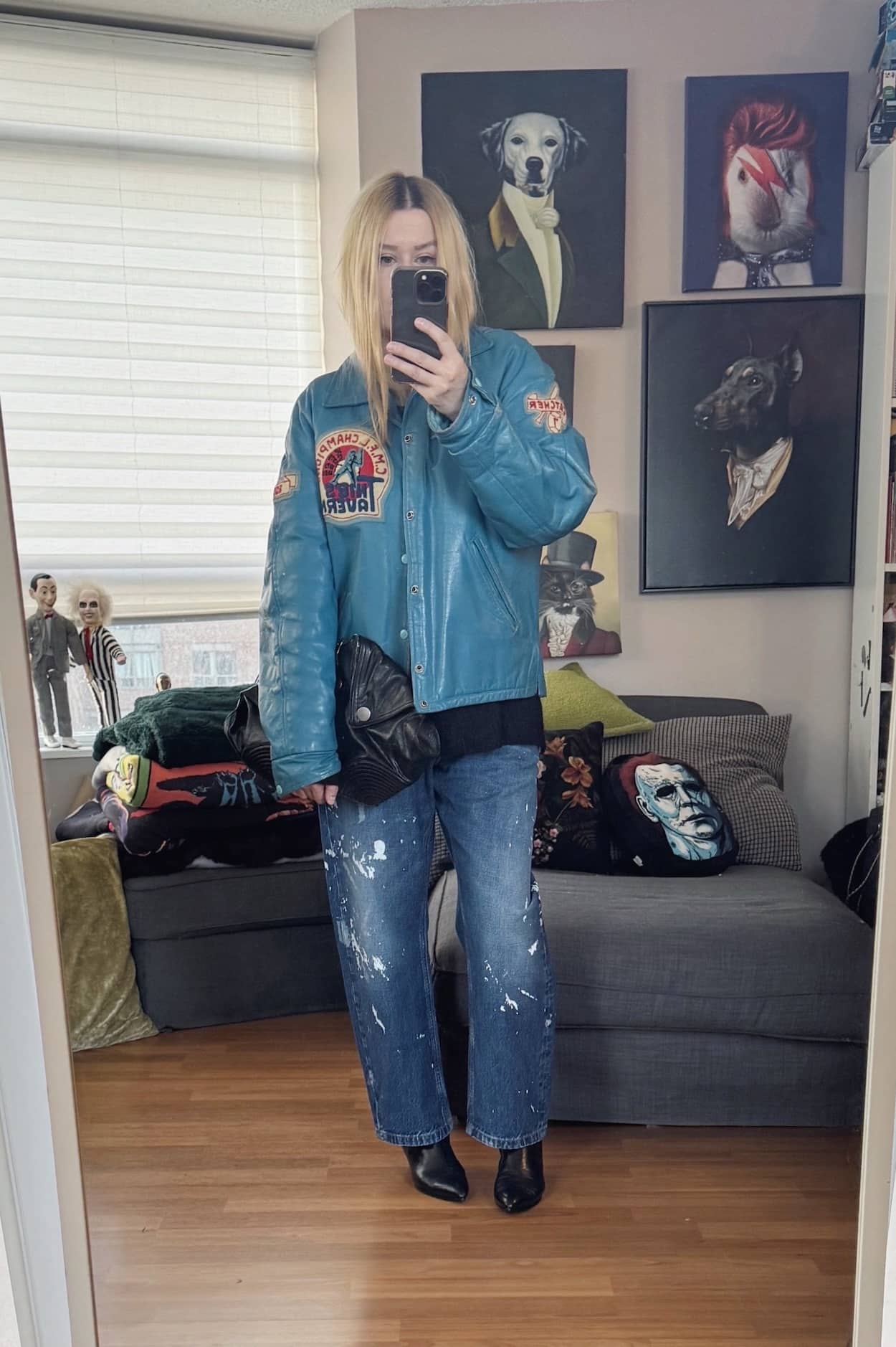 Sara of livelovesara is standing in front of a mirror taking a photo of her outfit. She is wearing a blue vintage leather jacket, paint splatter jeans, black boots, a black sweater, and an Alexander McQueen de manta clutch.