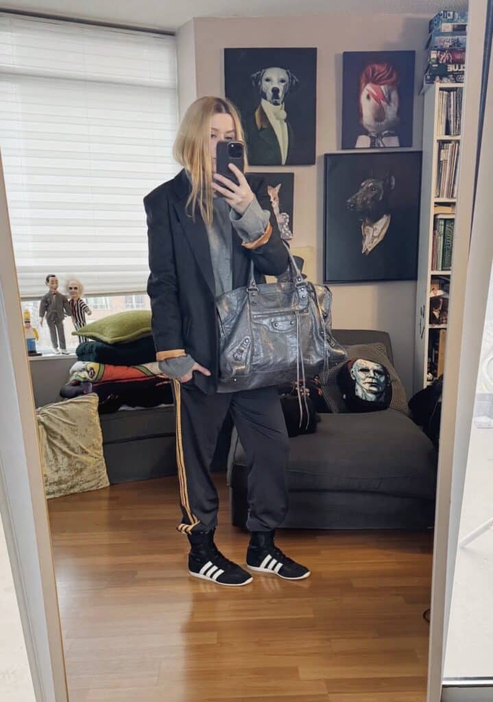 Sara of livelovesara is standing in front of a mirror taking a photo of her outfit. She is wearing a grey turtleneck sweater under a grey blazer, adidas track pants, Adidas wrestling sneakers, and a large Balenciaga city work bag.