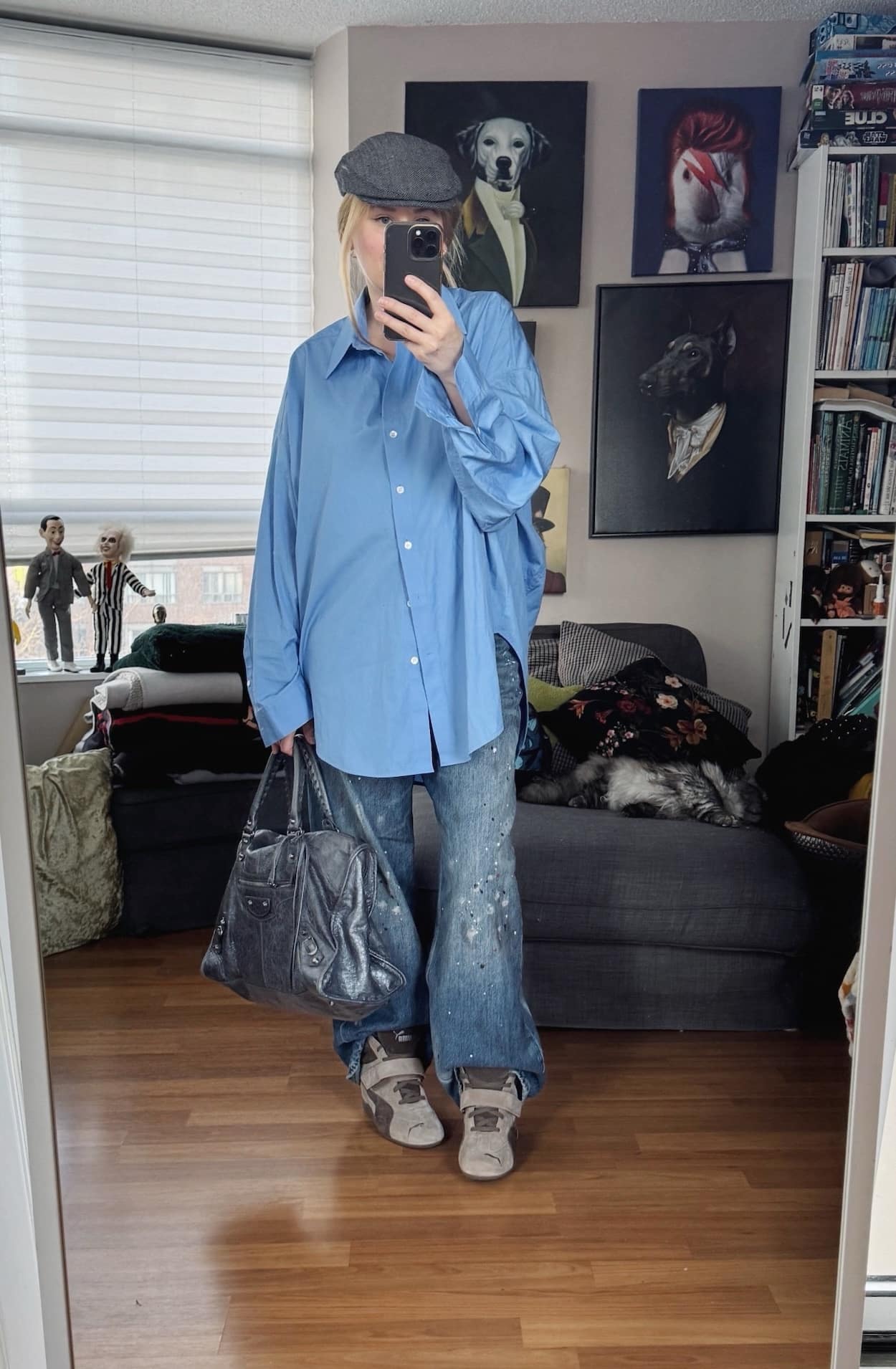 Sara of livelovesara is standing in front of a mirror taking a photo of her outfit. She is wearing an oversized blue button up over paint splattered jeans, wedge sneakers, a grey driving cap, and a grey Balenciaga bag.