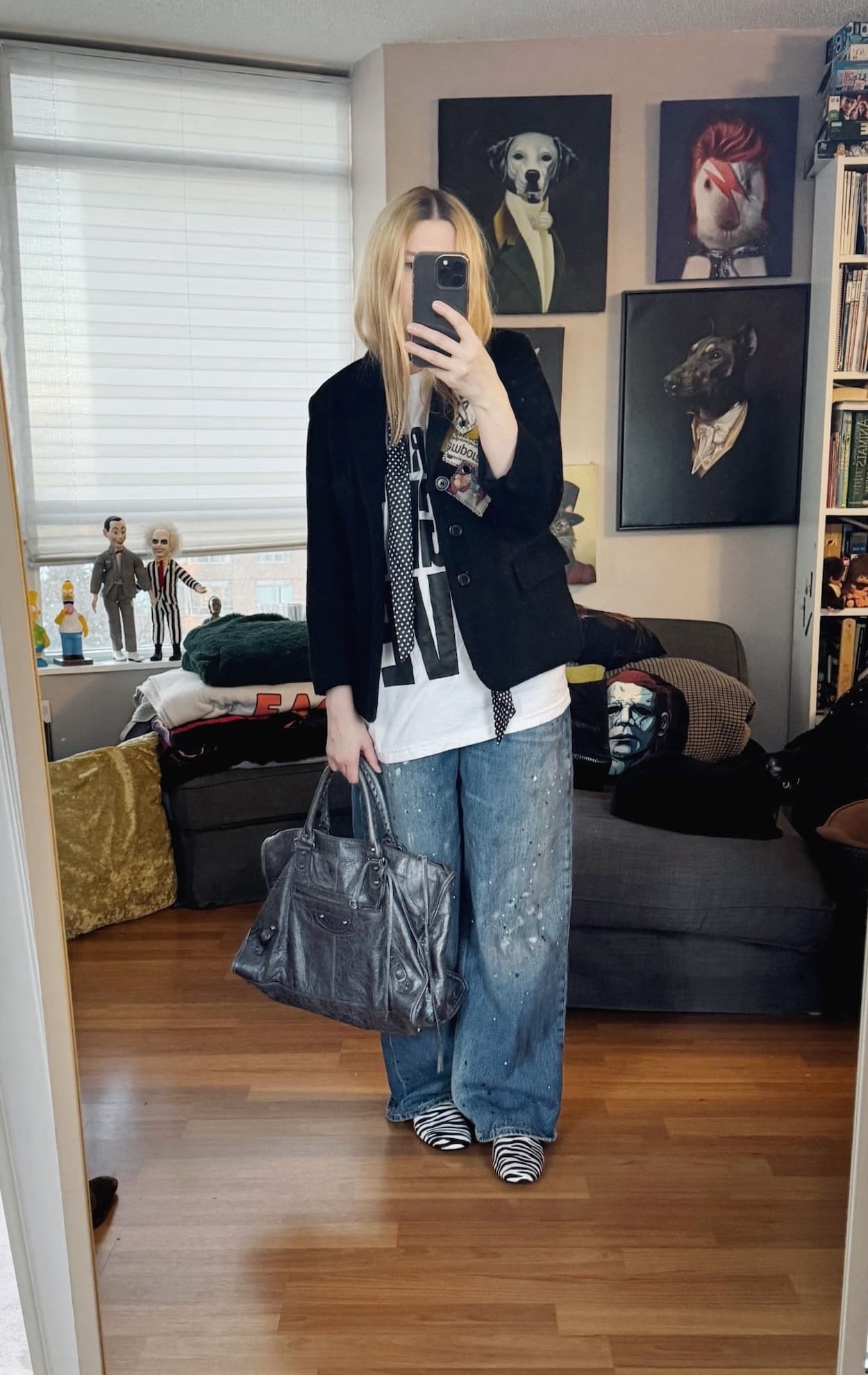 Sara of livelovesara is standing in front of a mirror taking a photo of her outfit. She is wearing a white t-shirt with Saturday Night Live in black large letters. Over that is a black vintage blazer, polka dot skinny scarf, paint splattered, wide leg jeans, zebra print loafers, and a grey Balenciaga city work bag.