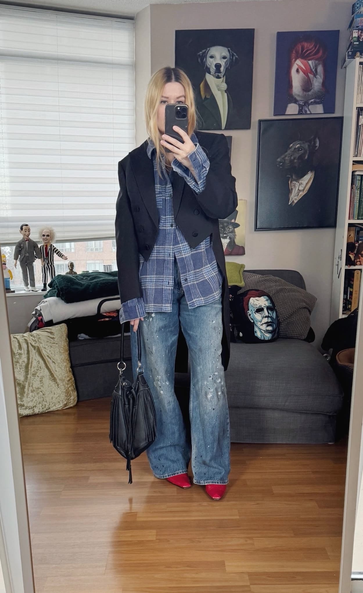 Sara of livelovesara is standing in front of a mirror taking a photo of her outfit. She is wearing a blue plaid, flannel shirt under a vintage black tailcoat, paint splatter jeans, red vintage boots, and is carrying a black, fringe bucket bag.