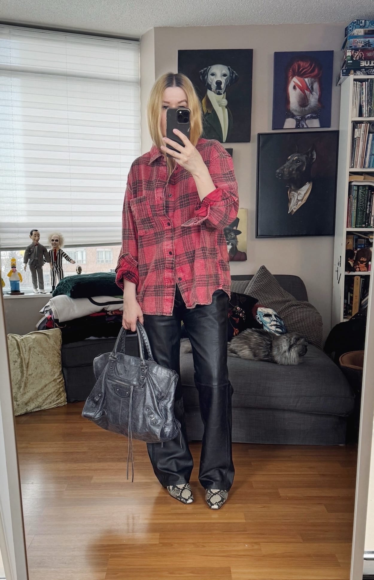 Sara of livelovesara is standing in front of a mirror taking a photo of her outfit. She is wearing a red and black plaid flannel shirt, with black vintage leather trousers, snake print boots, and is carrying a grey Balenciaga bag.