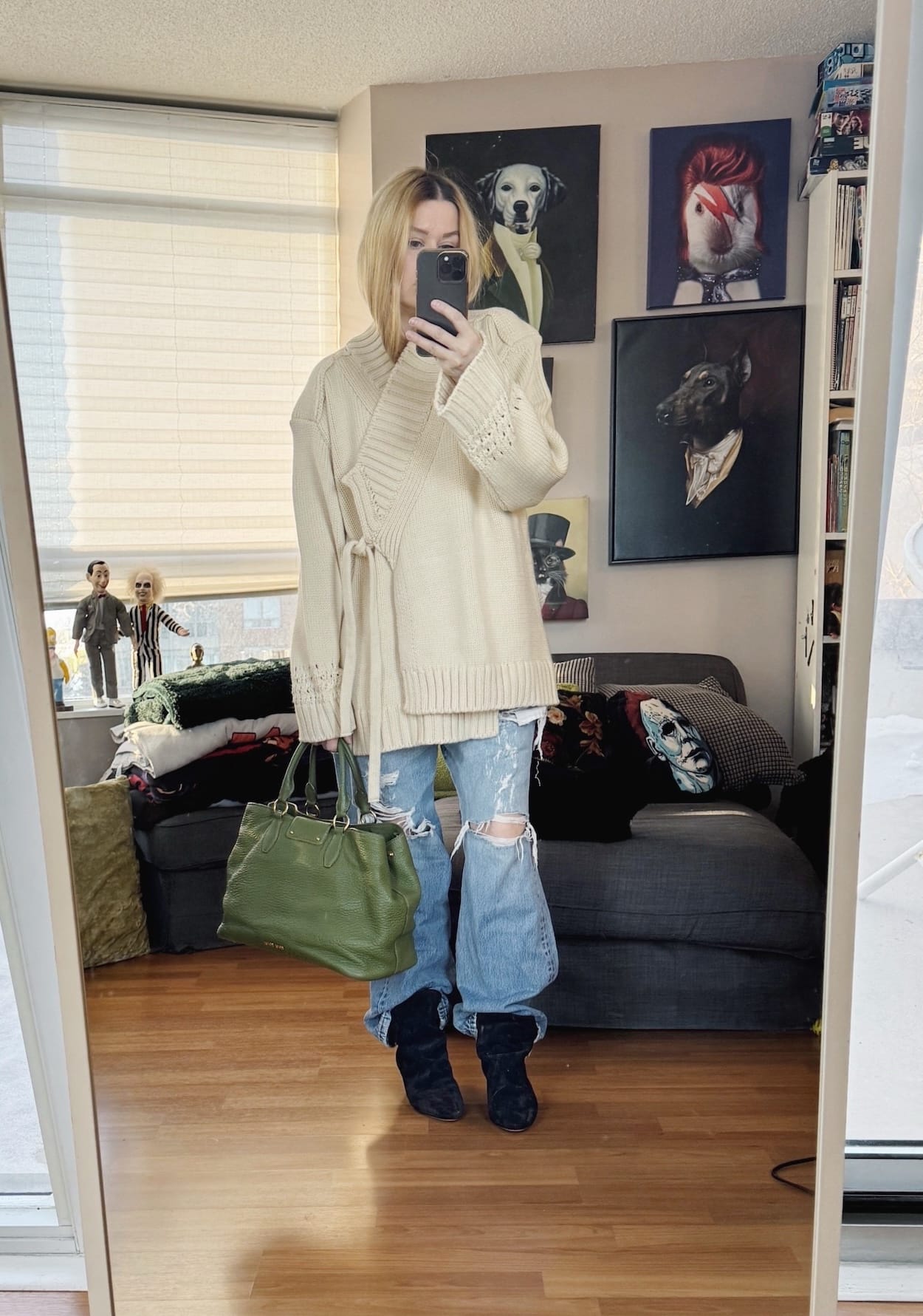 Sara of livelovesara is standing in front of a mirror taking a photo of her outfit. She is wearing a cream, oversized wrap cardigan over a white t-shirt, distressed vintage Levi's, black slouchy boots, and a green tote.
