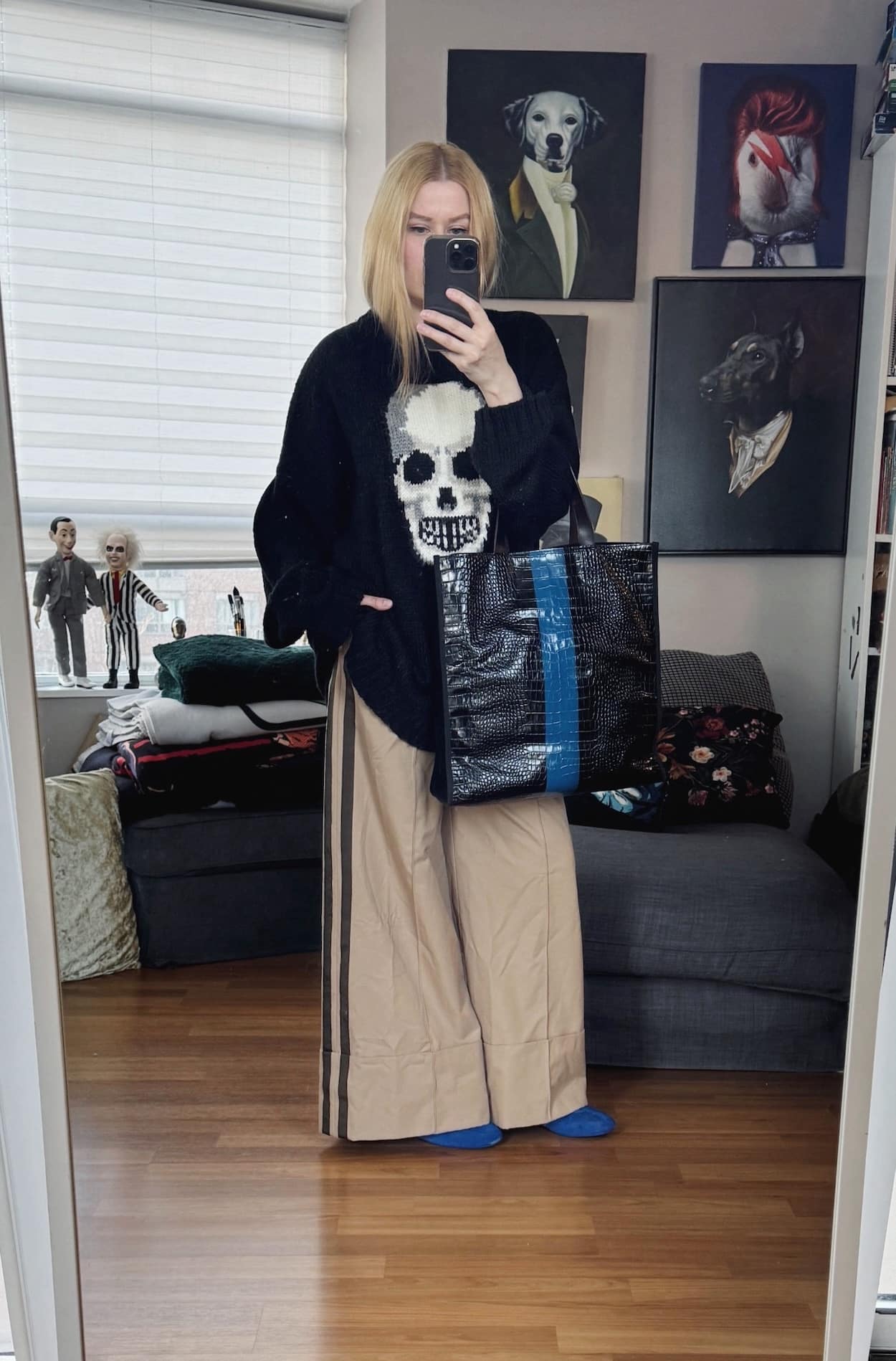 Sara of livelovesara is standing in front of a mirror taking a photo of her outfit. She is wearing a black sweater with a large skull on front, with wide leg tan trousers, blue Adidas Rasant sneakers, and a blue and black striped tote.