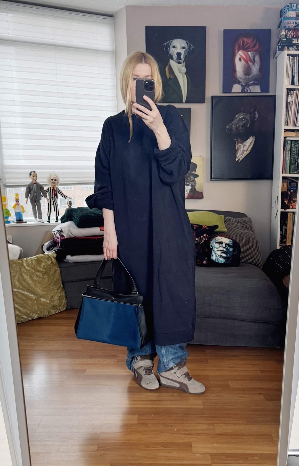 Sara of livelovesara is standing in front of a mirror taking a photo of her outfit. She is wearing a navy blue oversized sweatshirt dress over paint splatter jeans, Puma wedge sneakers, and a blue Celine edge bag.