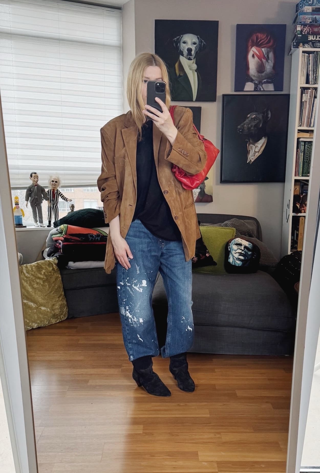 Sara of livelovesara is standing in front of a mirror taking a photo of her outfit. She is wearing a black t-shirt with paint splattered jeans, a brown suede blazer, black slouchy boots, and a red prada Y2K bag.