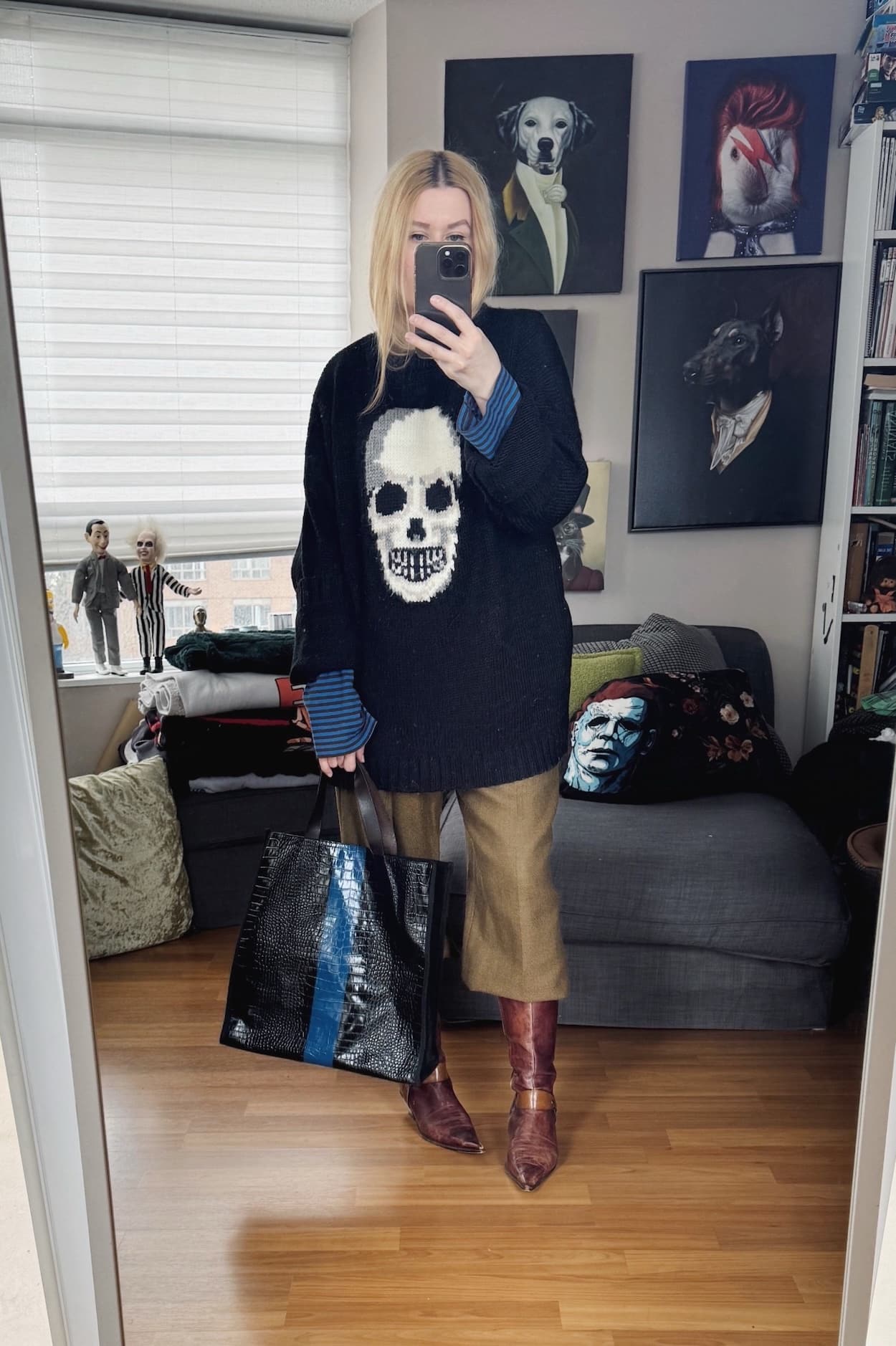 Sara of livelovesara is standing in front of a mirror taking a photo of her outfit. She is wearing a black sweater with a white skull over a blue and brown striped shirt, brown vintage trousers tucked into brown vintage boots, and a blue and black striped bag.