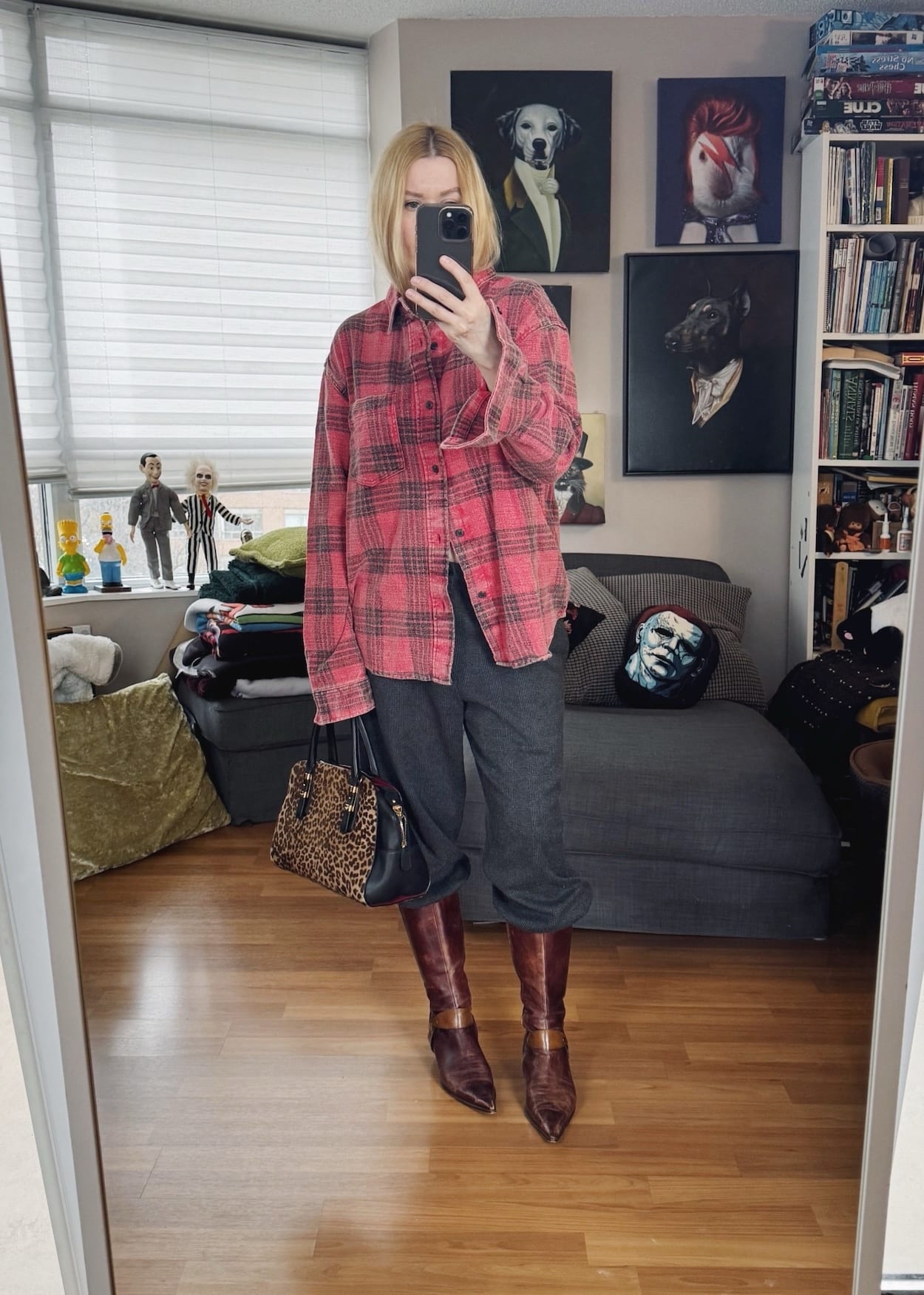 Sara of livelovesara is standing in front of a mirror taking a photo of her outfit. She is wearing a red plaid shirt, Grey trousers, vintage brown knee high boots, and animal print bag.
