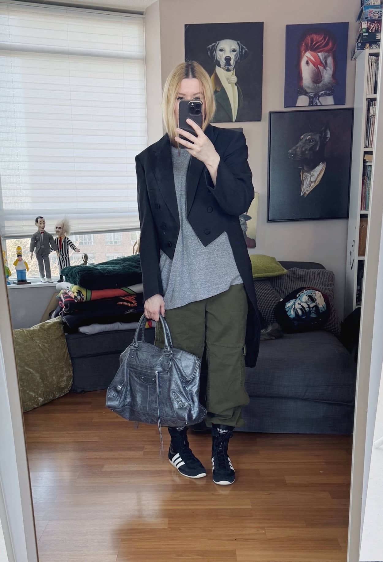 Sara of livelovesara is standing in front of a mirror taking a photo of her outfit. She is wearing a worn grey tee under a black vintage tailcoat, cargo pants, Adidas wrestling sneakers, and a Balenciaga city work bag