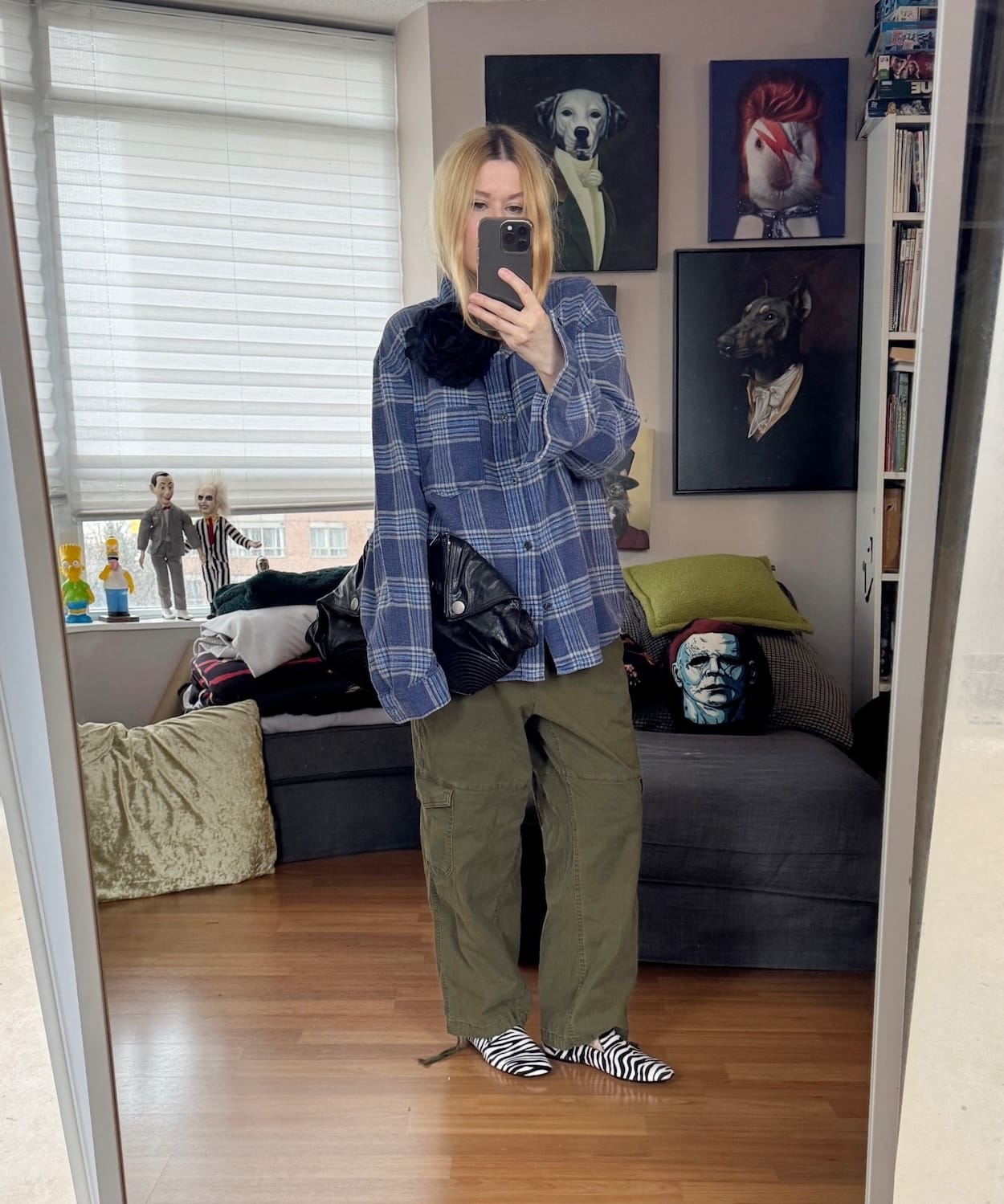 Sara of livelovesara is standing in front of a mirror taking a photo of her outfit. She is wearing a blue plaid shirt with olive cargo pants. zebra print loafers, a large black flower brooch, and is carrying an Alexander McQueen clutch.