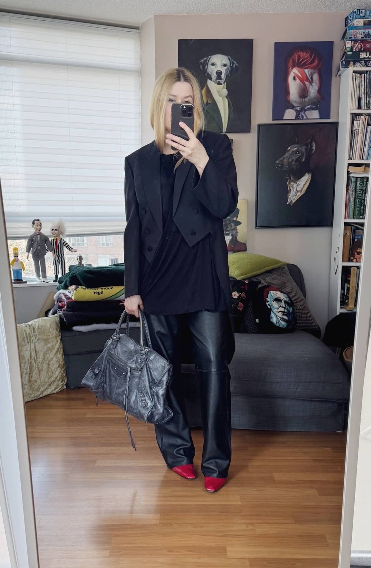Sara of livelovesara is standing in front of a mirror taking a photo of her outfit. She is wearing a black t-shirt with black vintage leather pants, a vintage tailcoat, red boots, and a Balenciaga work bag.