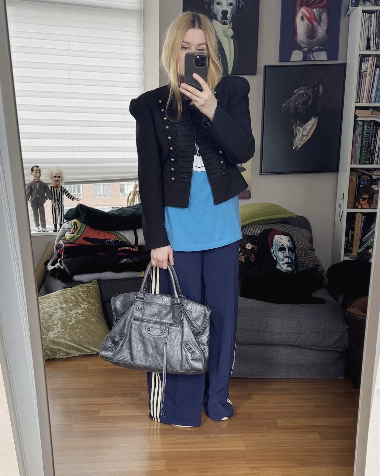 Sara of livelovesara is standing in front of a mirror taking a photo of her outfit. She is wearing a SOnic Youth tee under a Napoleon jacket, with blue Adidas wide leg track pants, Adidas black boxing sneakers, and a grey Balenciaga work bag.