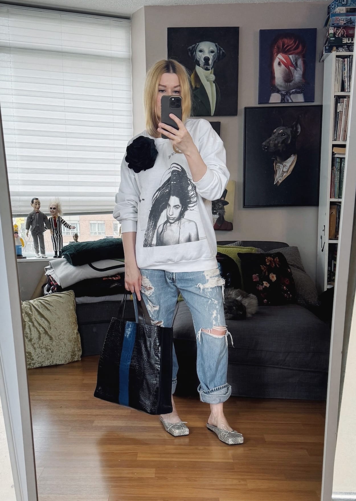 Sara of livelovesara is standing in front of a mirror taking a photo of her outfit. She is wearing a white PJ Harvey sweatshirt with distressed vintage Levi's, snake print mules, a large black flower brooch, and is carrying a black and blue striped tote.